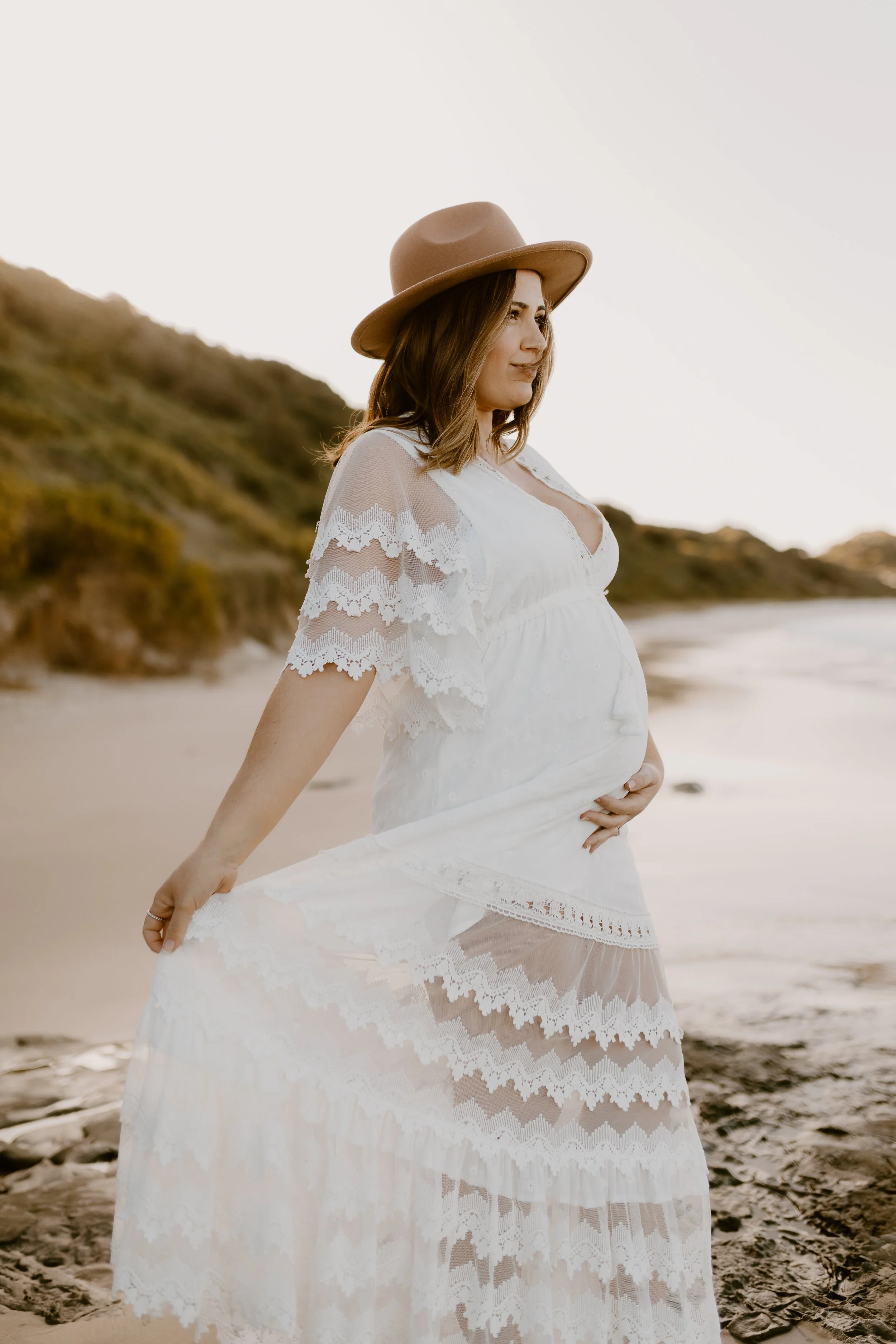 Pregnant woman in a white lace dress and a tan hat standing on a beach during sunset.