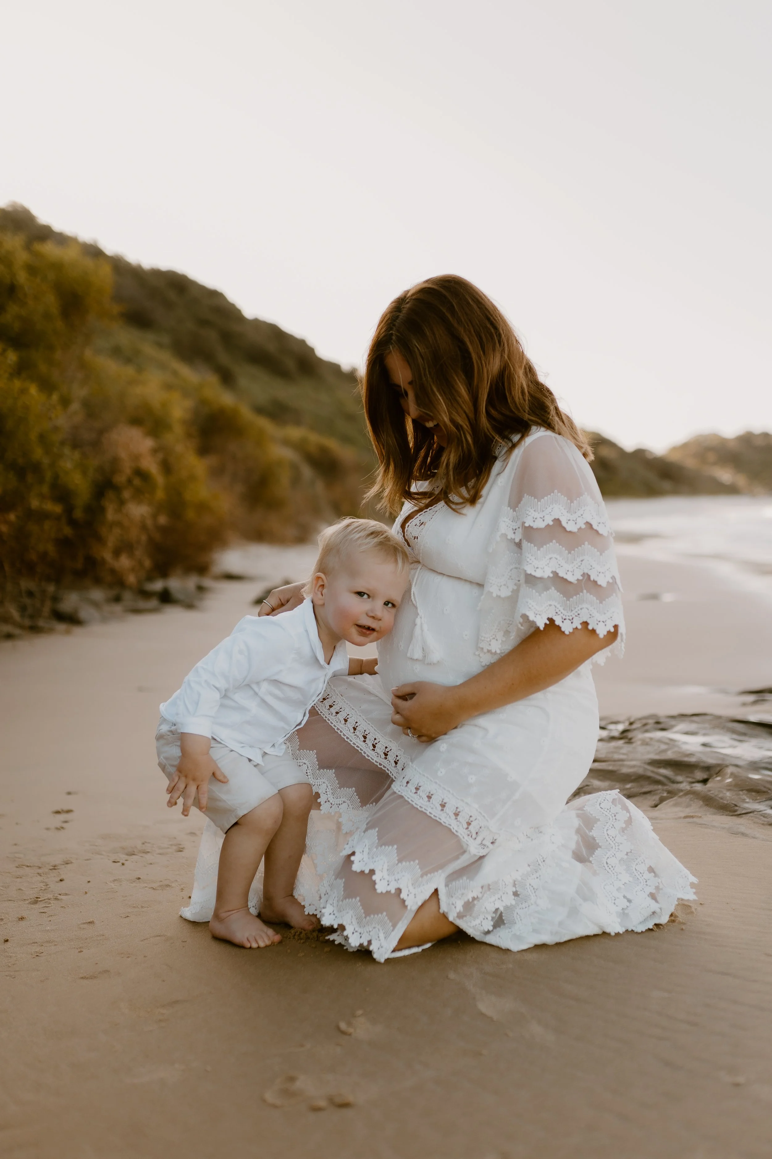 A pregnant woman and a young child on a beach during sunset, both dressed in white, with the woman kneeling and cradling her belly while the child leans in smiling.