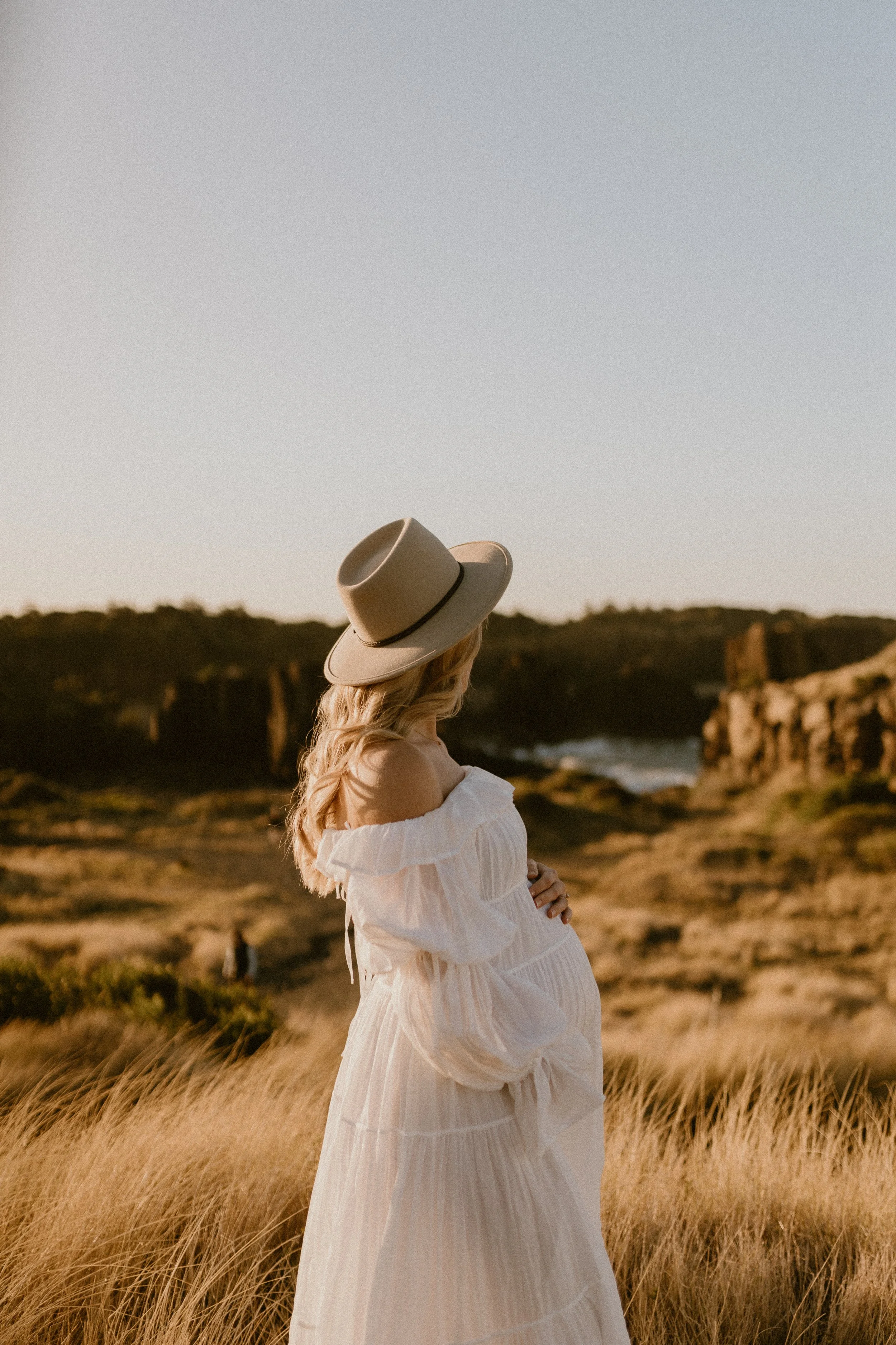 Pregnant woman with long blonde hair wearing a white off-shoulder dress and a beige hat standing in a field of tall grass during sunset.