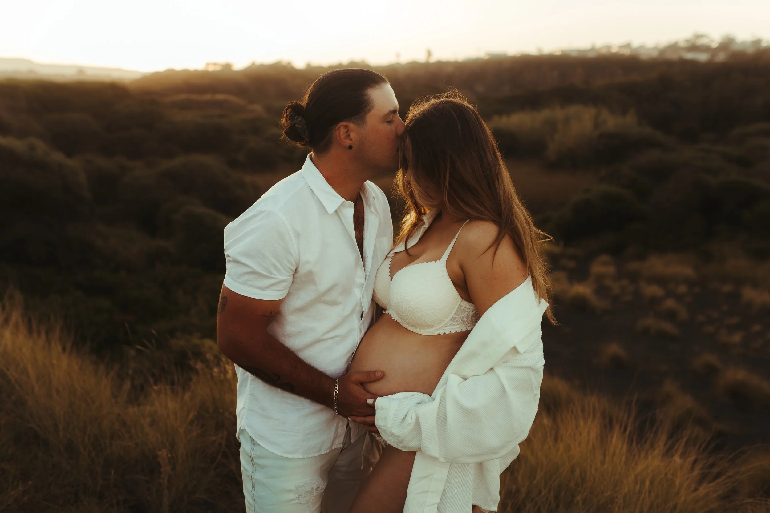 A pregnant woman and a man are standing outdoors in a grassy field during sunset, sharing a kiss.