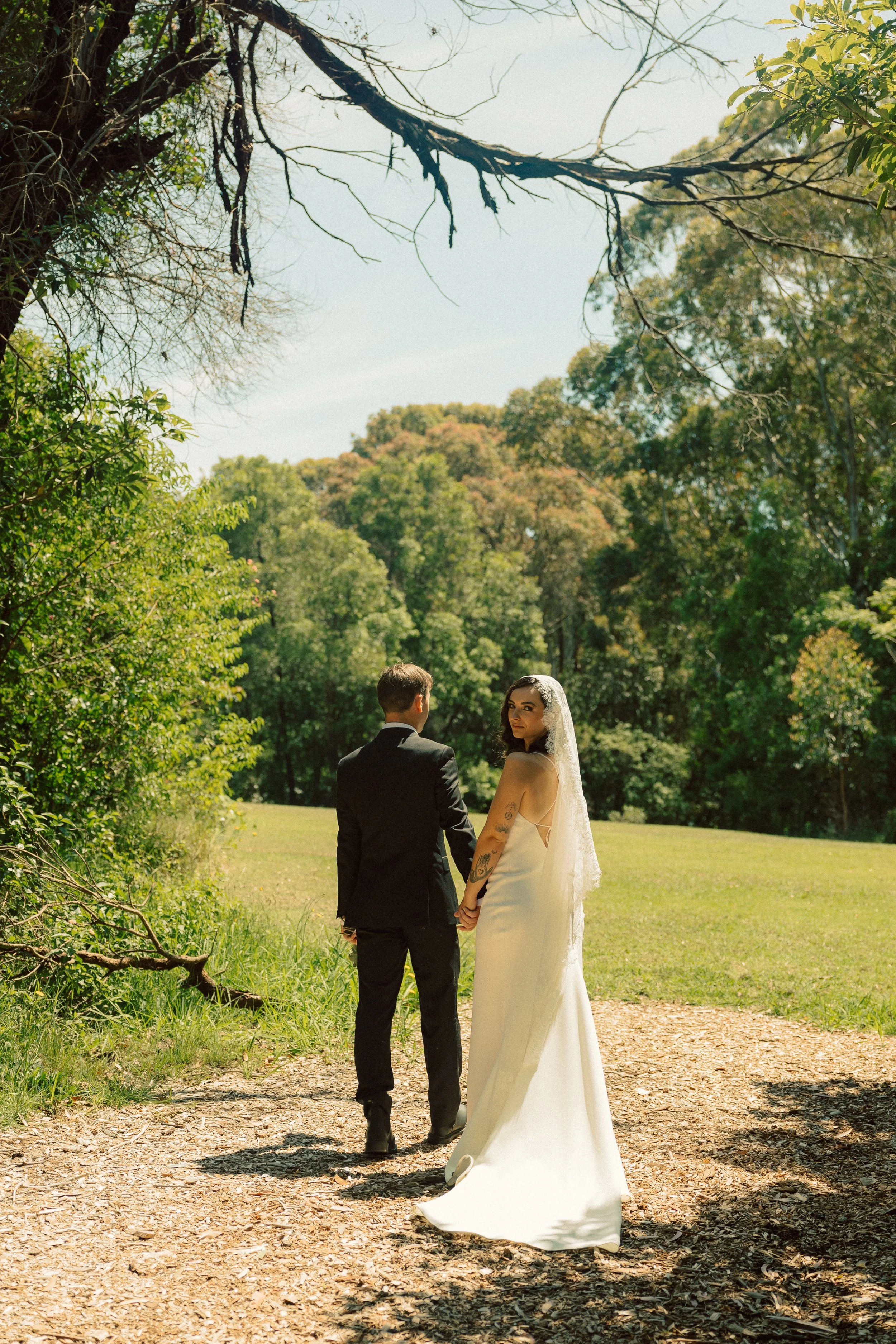 A couple dressed in wedding attire holding hands and walking along a wooded path in a park-like setting on a sunny day.