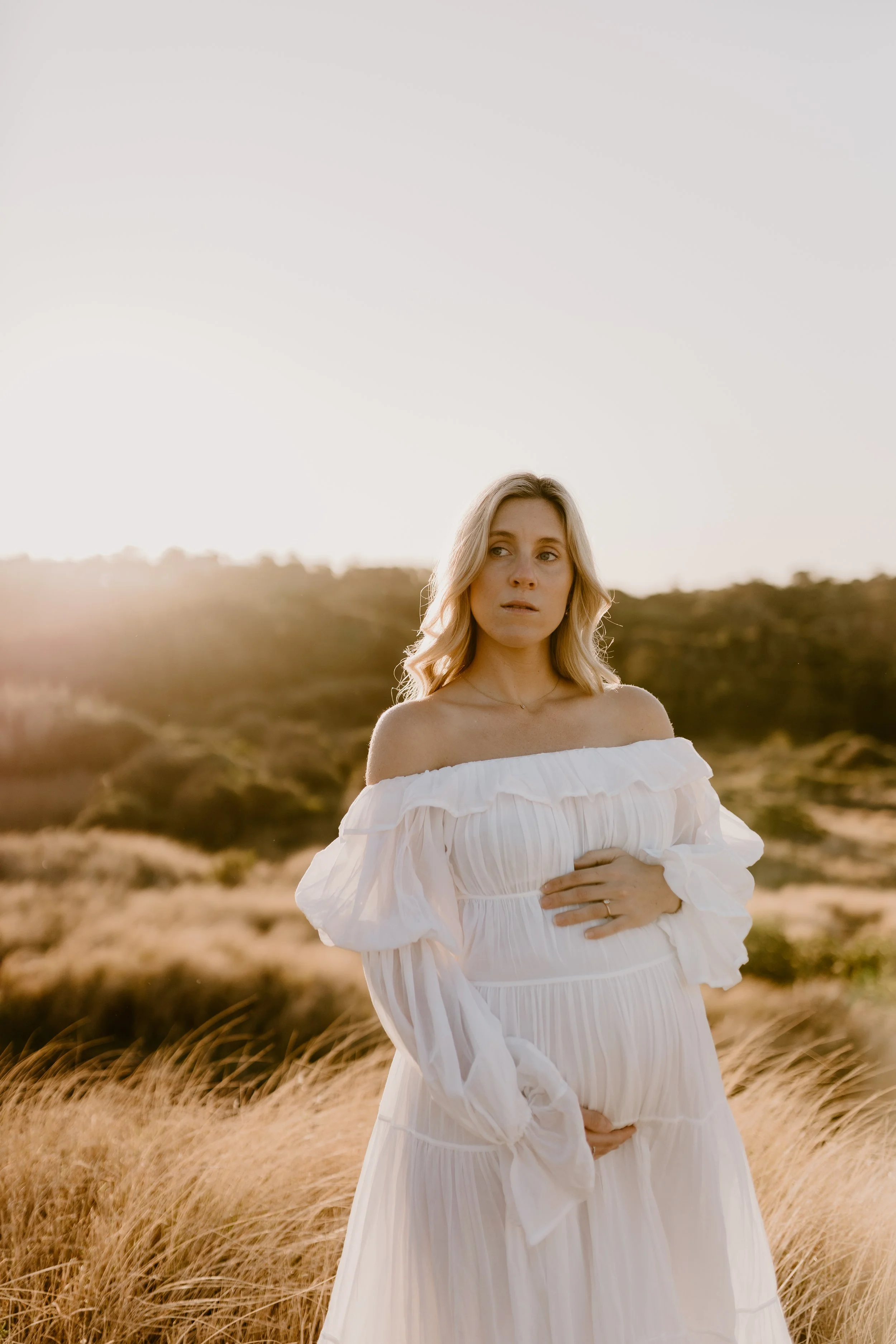 A pregnant woman wearing a white off-shoulder dress standing outdoors in a grassy field during sunset.