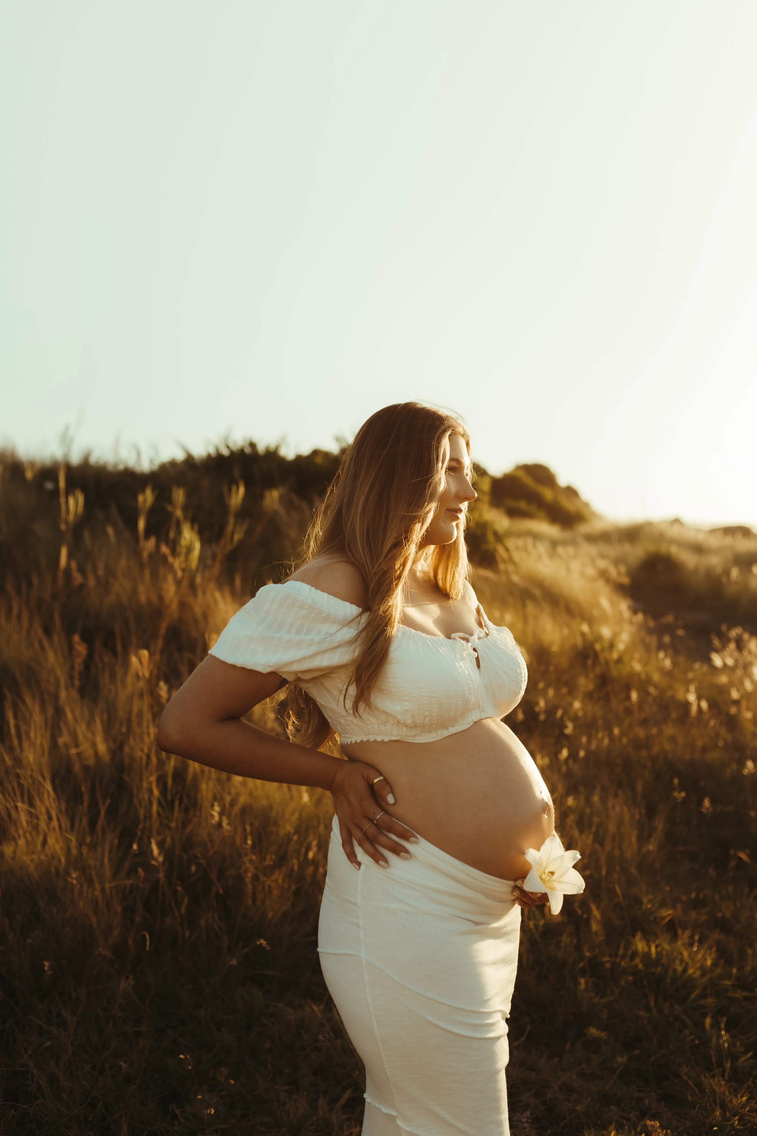 Pregnant woman in cream-colored dress holding her belly outdoors in a grassy area during sunset.