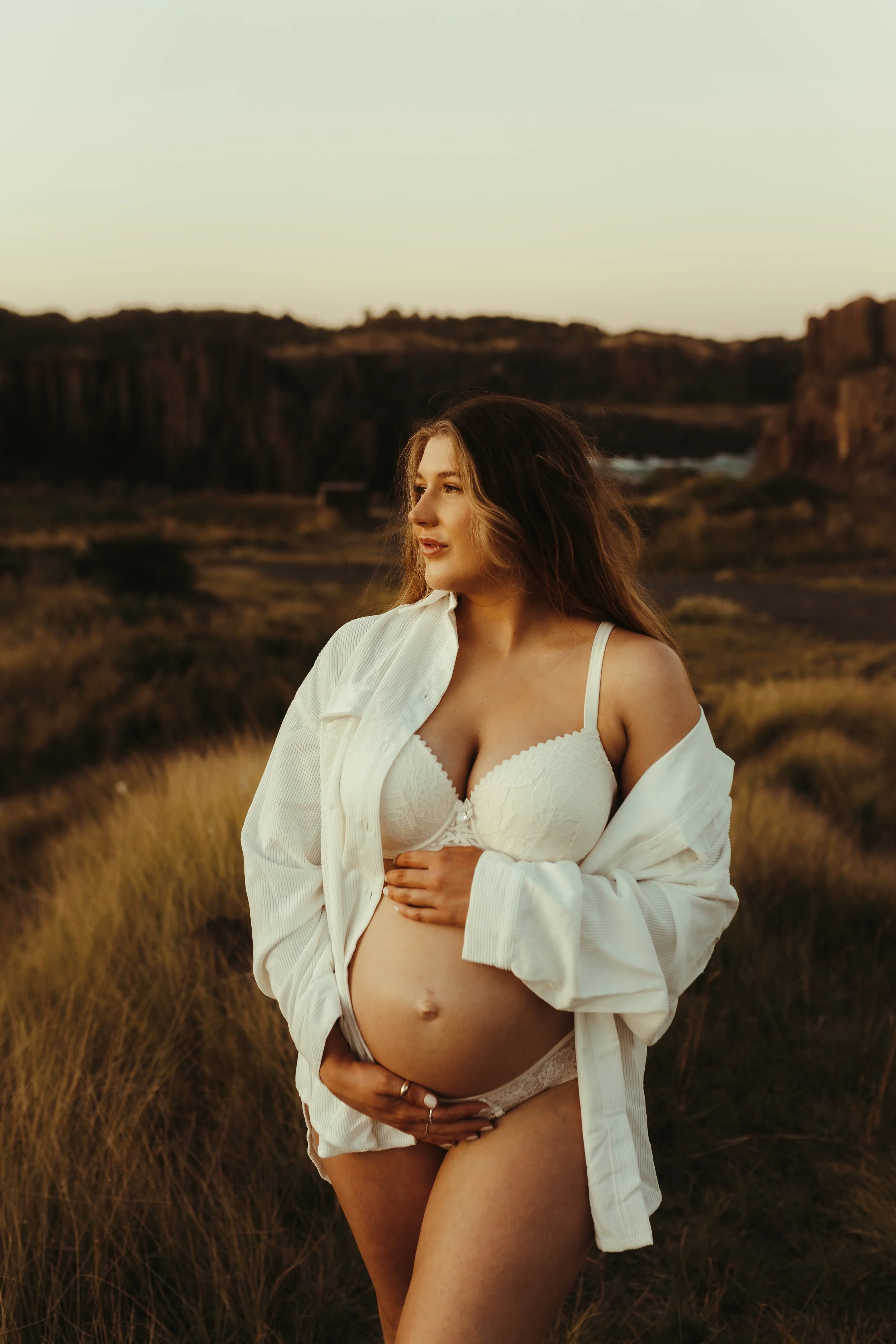 A pregnant woman in white lingerie and an open shirt stands in a grassy field at sunset, with rocky cliffs in the background.