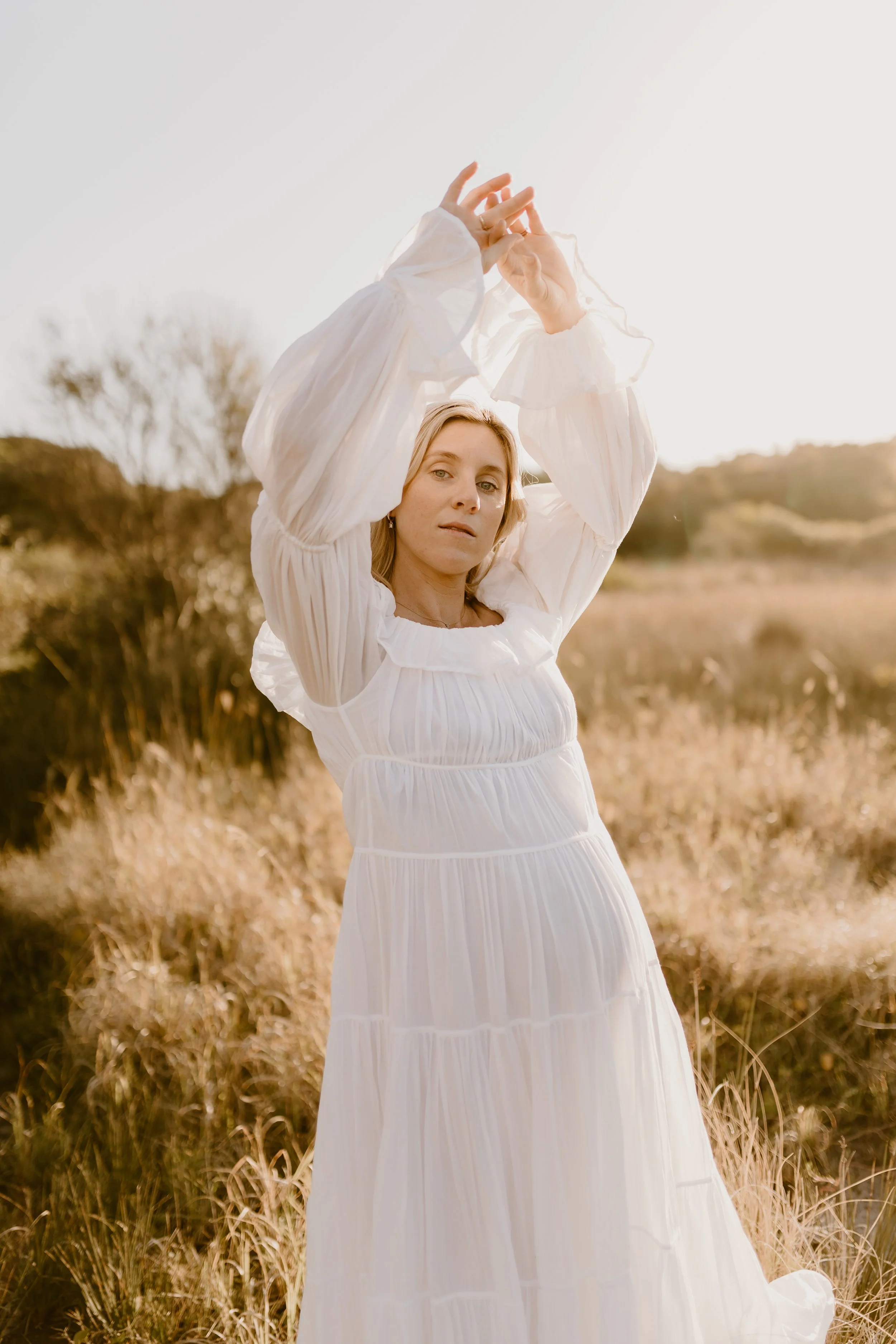 A woman in a white dress stands outdoors with her arms raised, in a natural landscape during golden hour.