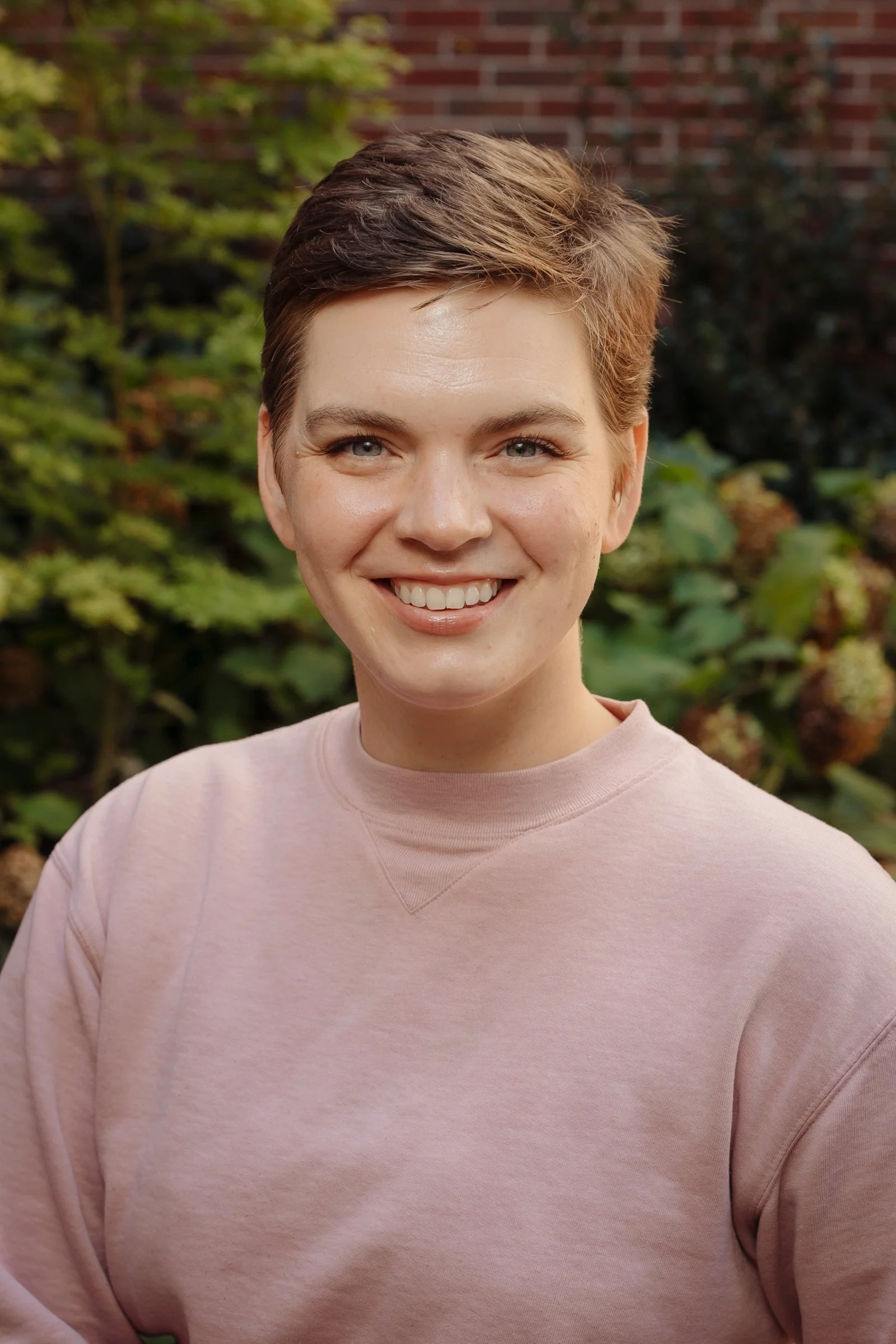 A young person with short brown hair smiling outdoors, wearing a light pink sweatshirt, with greenery and a brick wall in the background.