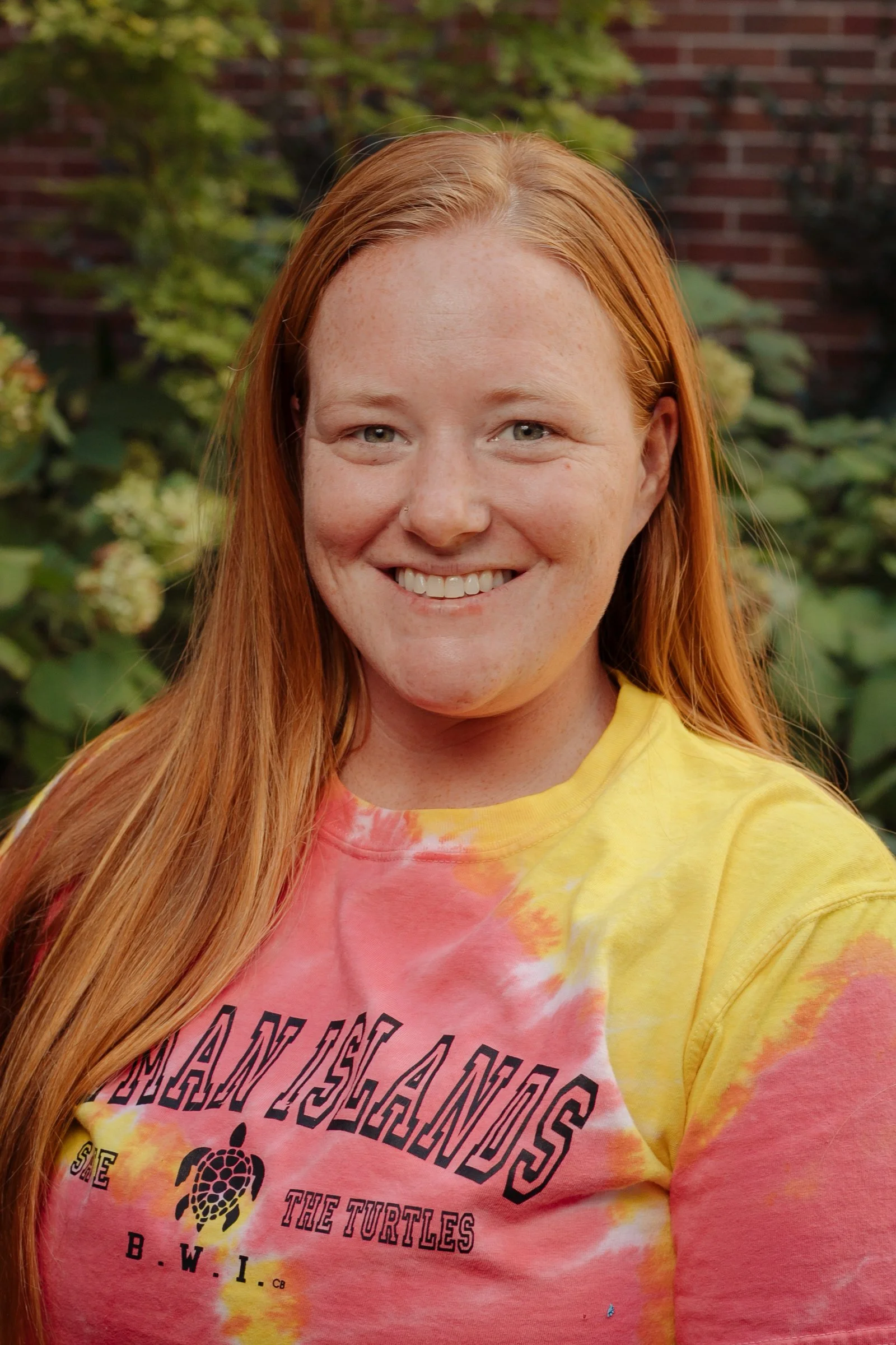 A woman with long red hair smiling outdoors in front of green foliage and a brick wall, wearing a tie-dye yellow and pink shirt that says 'SANDALS' and 'THE TURTLES'.