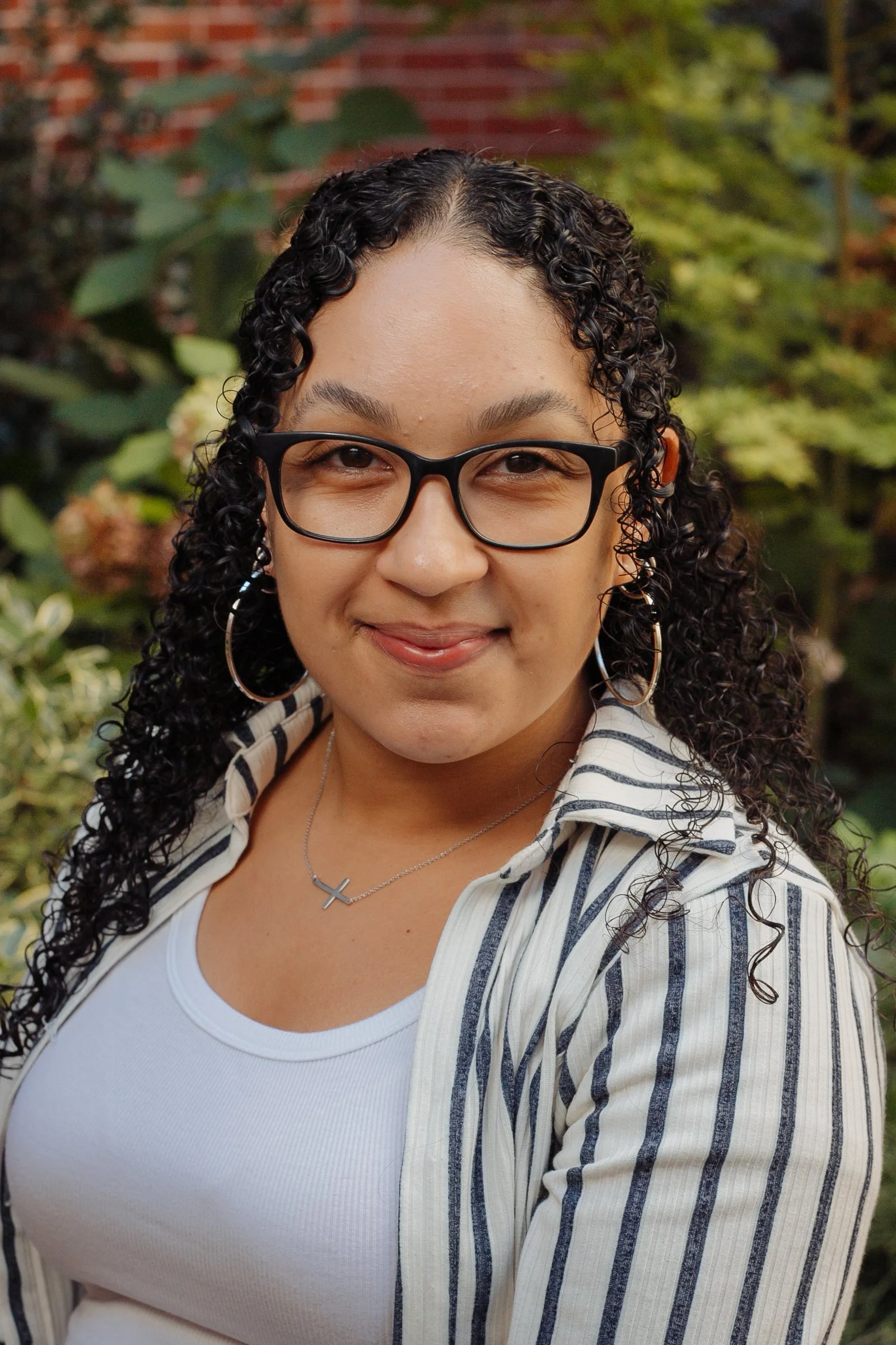 A woman with curly hair, glasses, hoop earrings, and a cross necklace, smiling outdoors with greenery and a brick wall in the background.