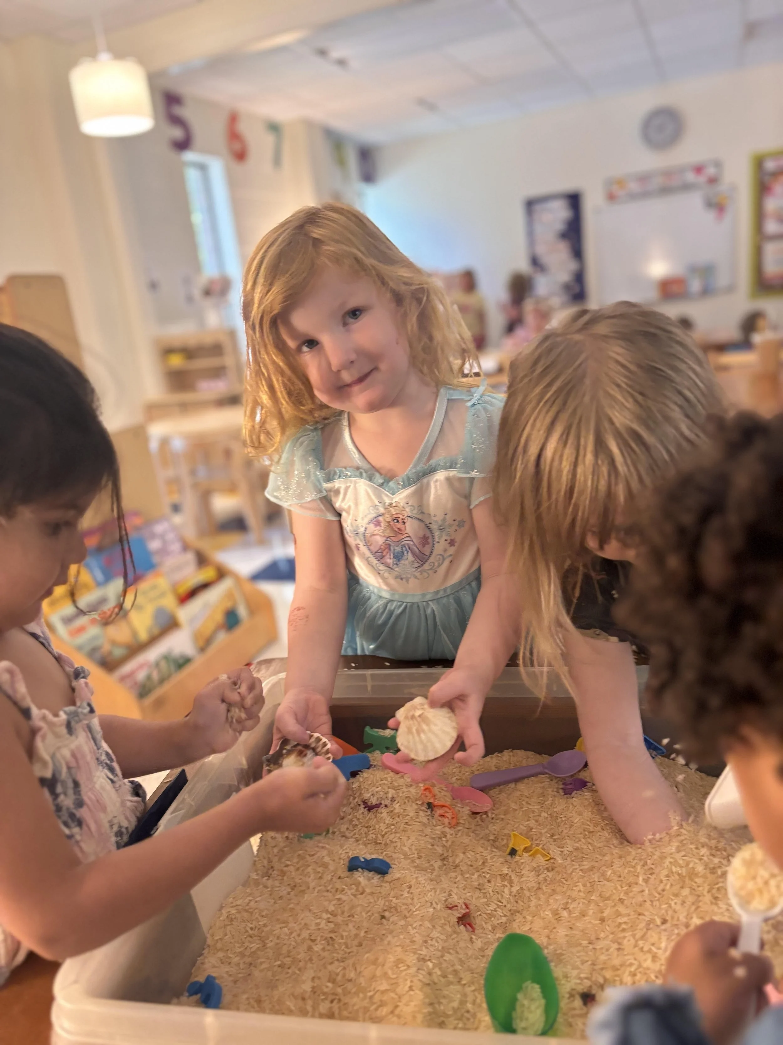 Children playing in a sandbox with seashells and plastic toys in a classroom decorated with numbers and colorful posters.