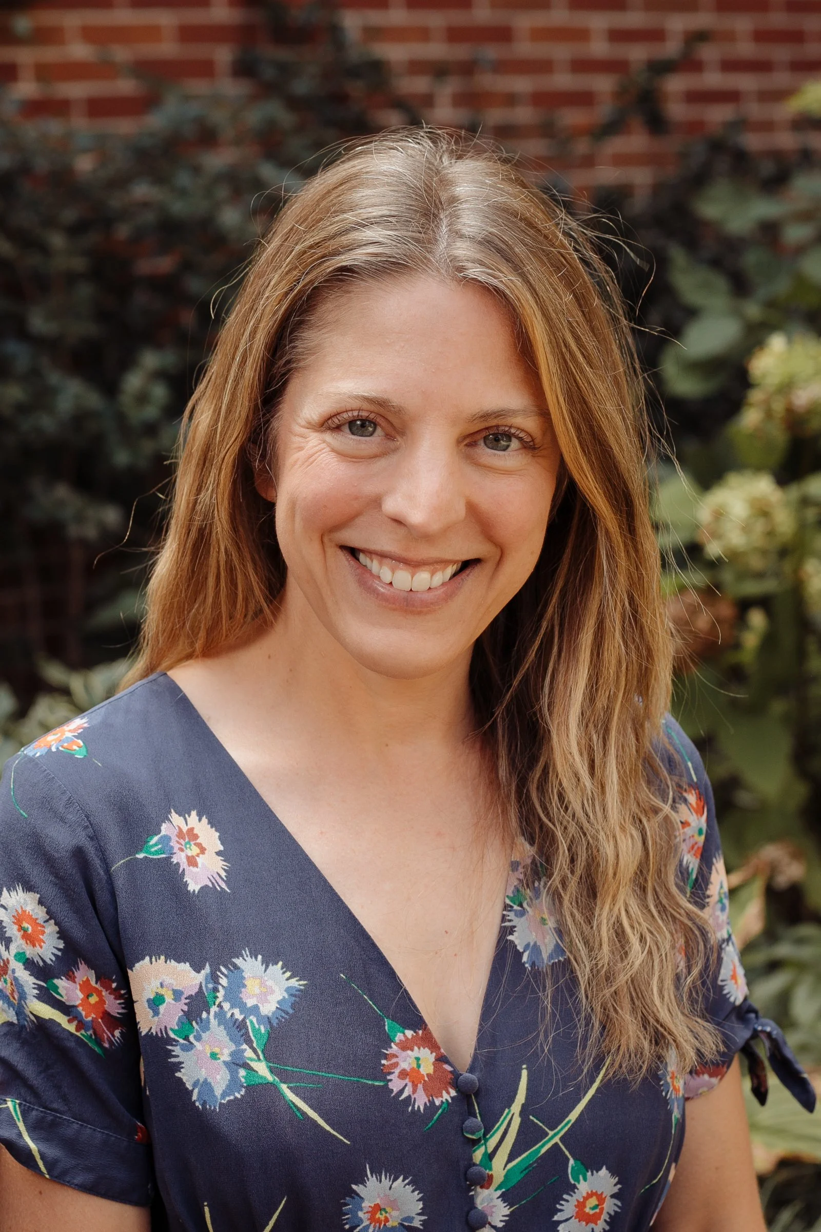 A woman with long, wavy light brown hair and blue eyes smiling outdoors, wearing a navy blue floral dress with short sleeves and buttons, with greenery and a brick wall in the background.