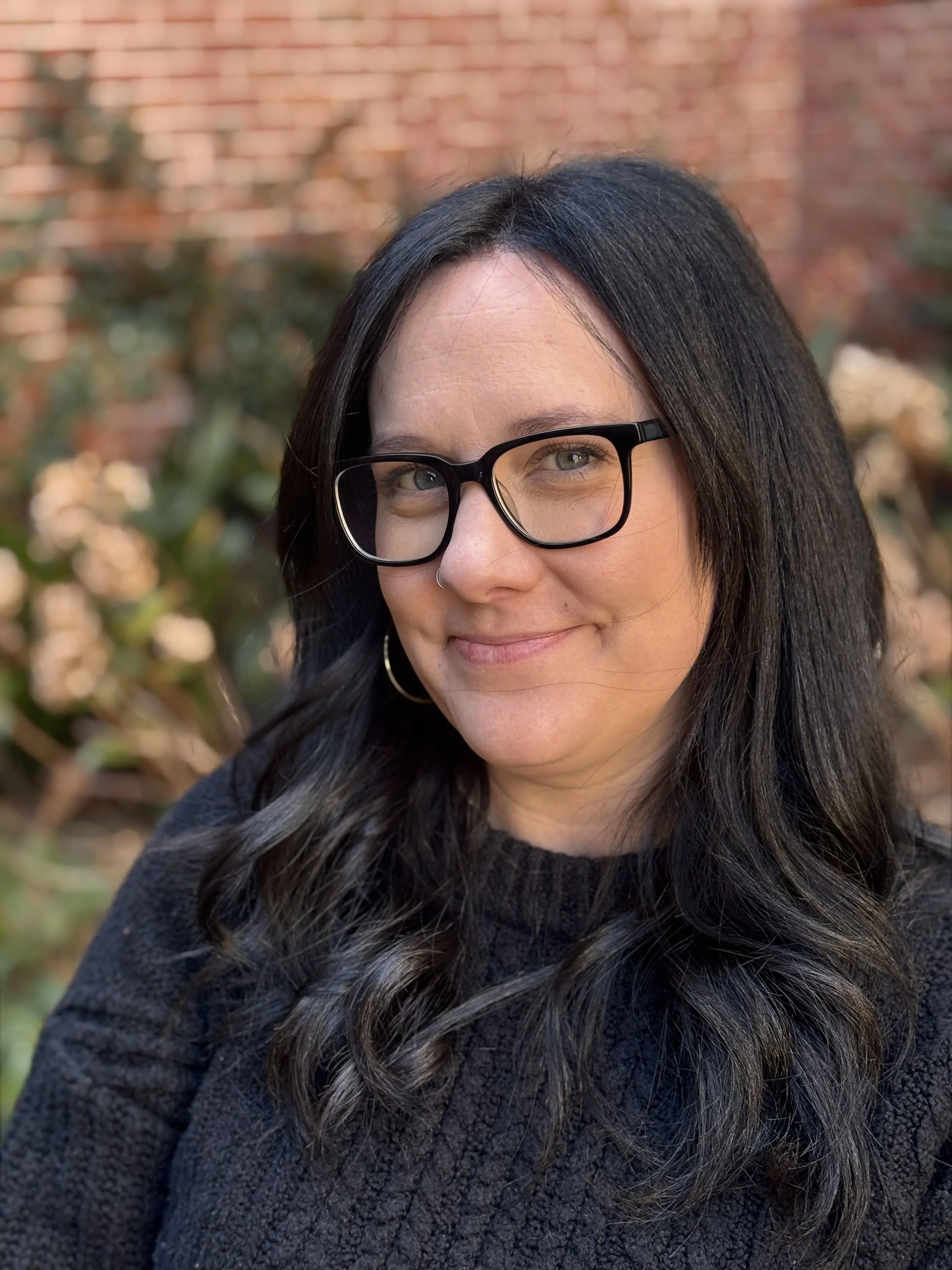Portrait of a woman with black hair, glasses, and a nose ring, smiling outdoors with blurred greenery and brick wall in the background.