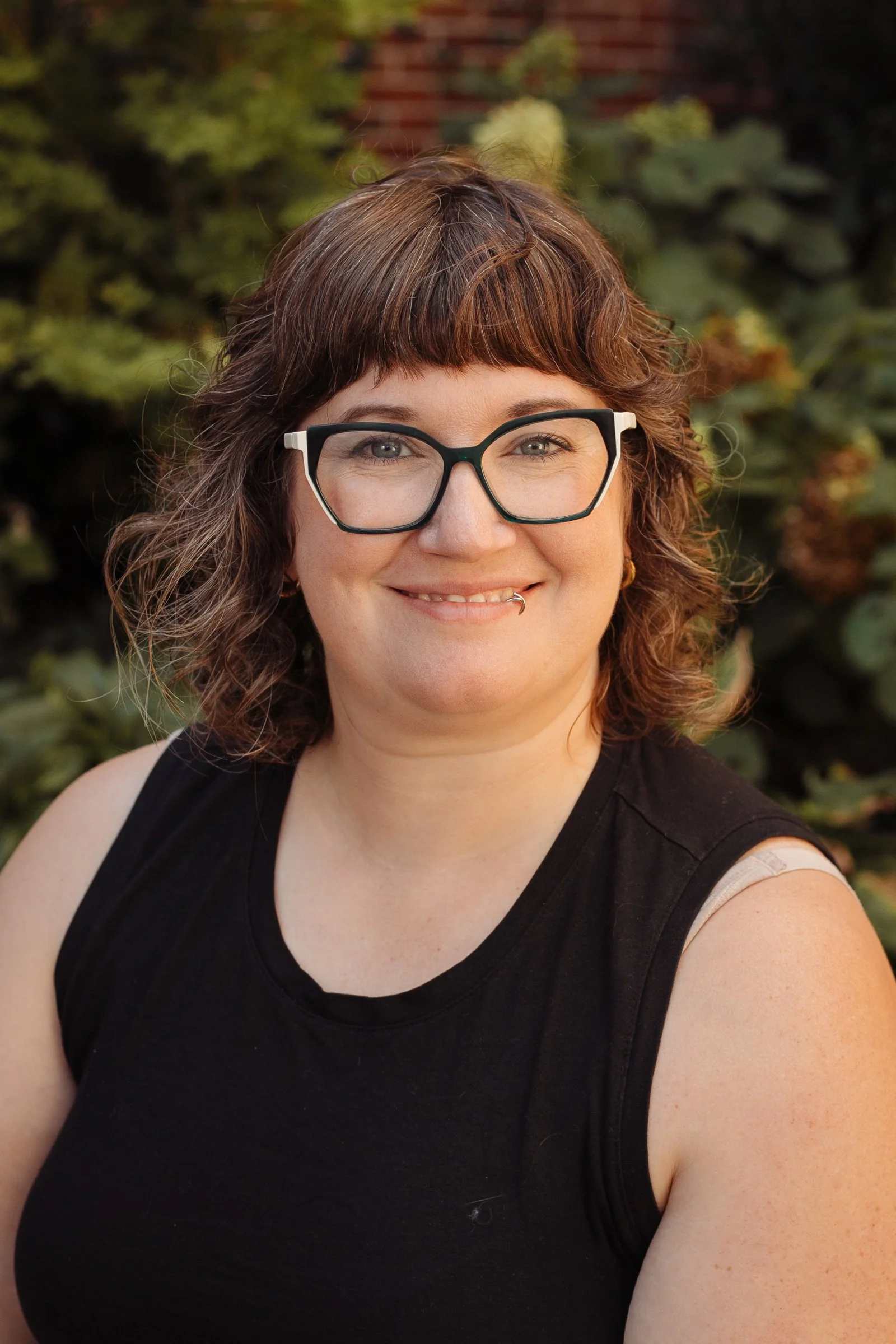 A woman with glasses and a lip ring smiling outdoors, wearing a black sleeveless top, with greenery in the background.