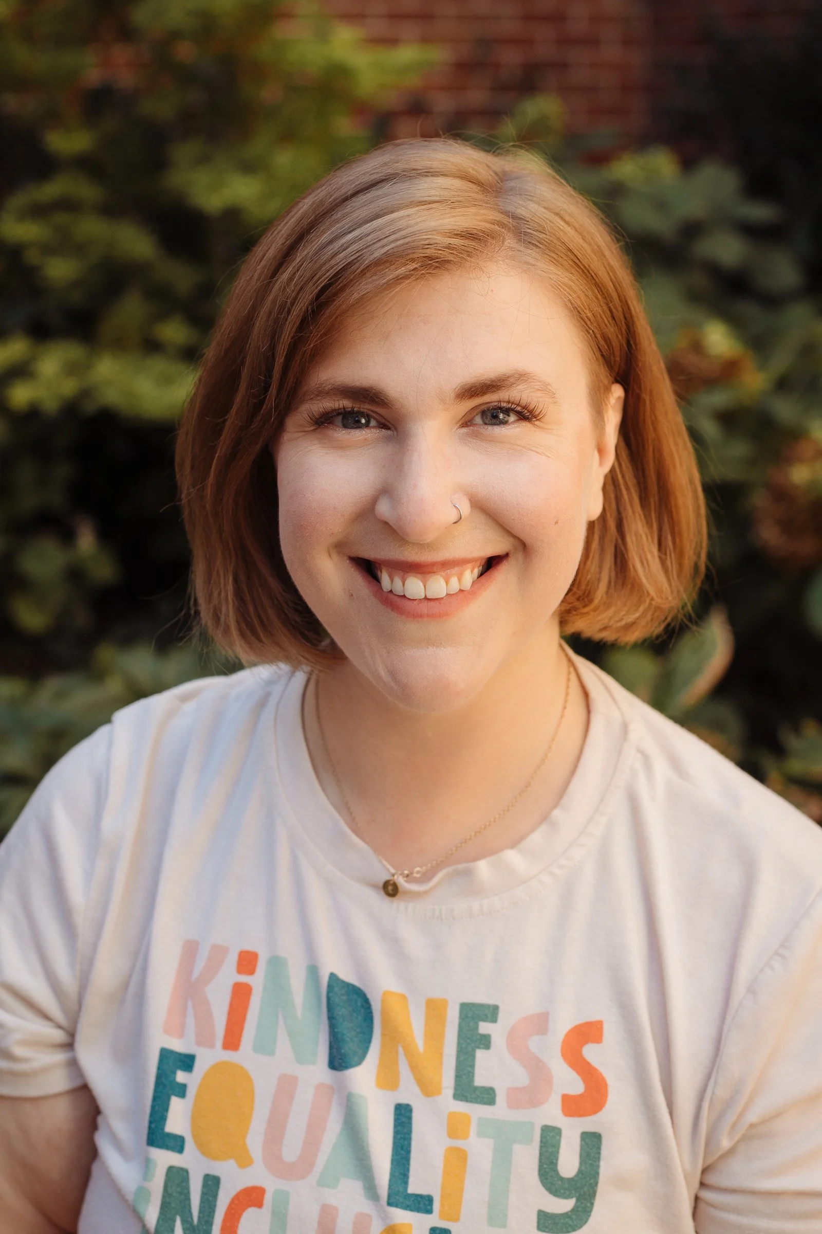 A smiling woman with shoulder-length red hair, wearing a white T-shirt with colorful text that reads "KINDNESS EQUALITY" and a small necklace, outdoors with greenery in the background.