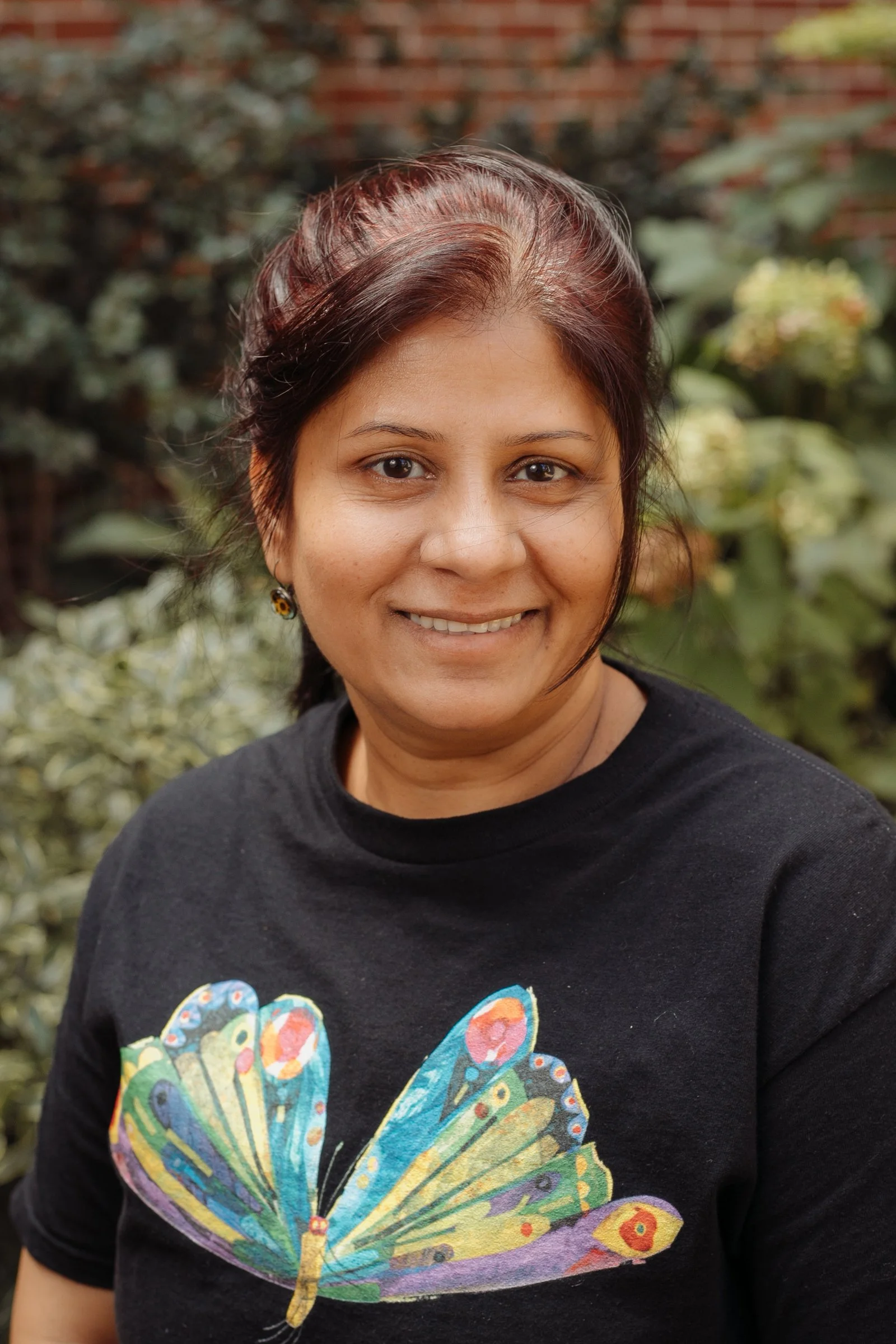 Portrait of a smiling woman with dark hair and earrings, wearing a black t-shirt with a colorful butterfly design, outdoors with green foliage in the background.
