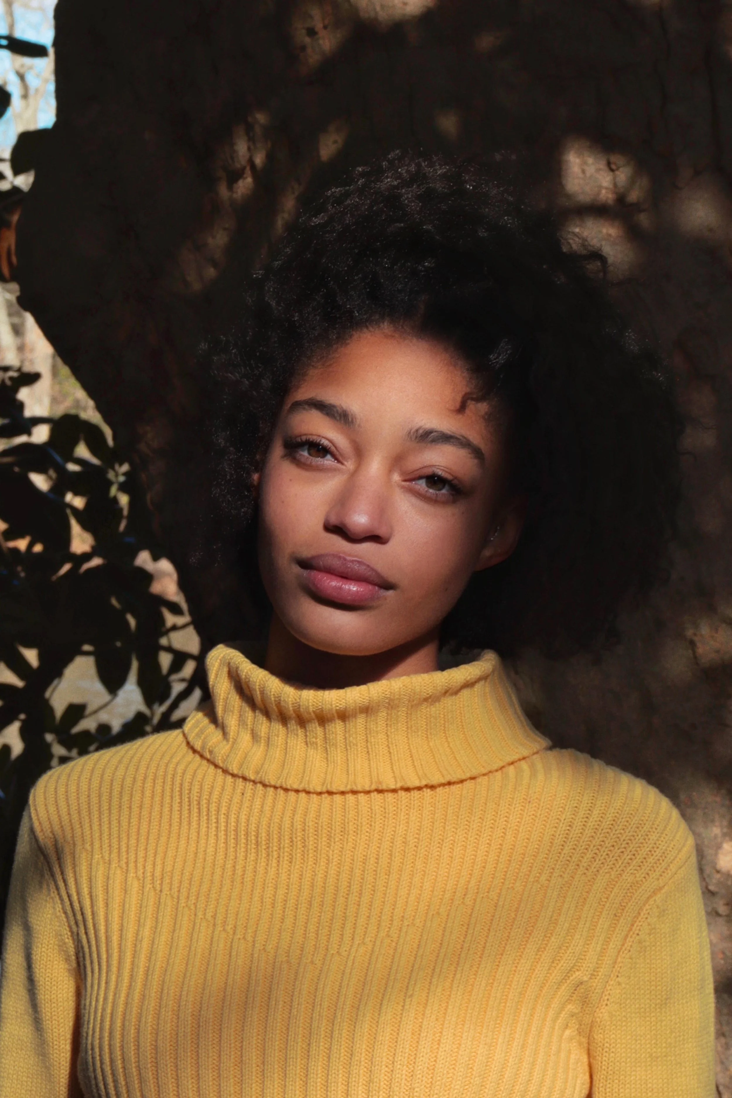 Infinit, a Black woman with dark natural hair, smiles gently in front of a shadowy outdoor setting. She wears a yellow turtleneck and their head is tilted to one side.