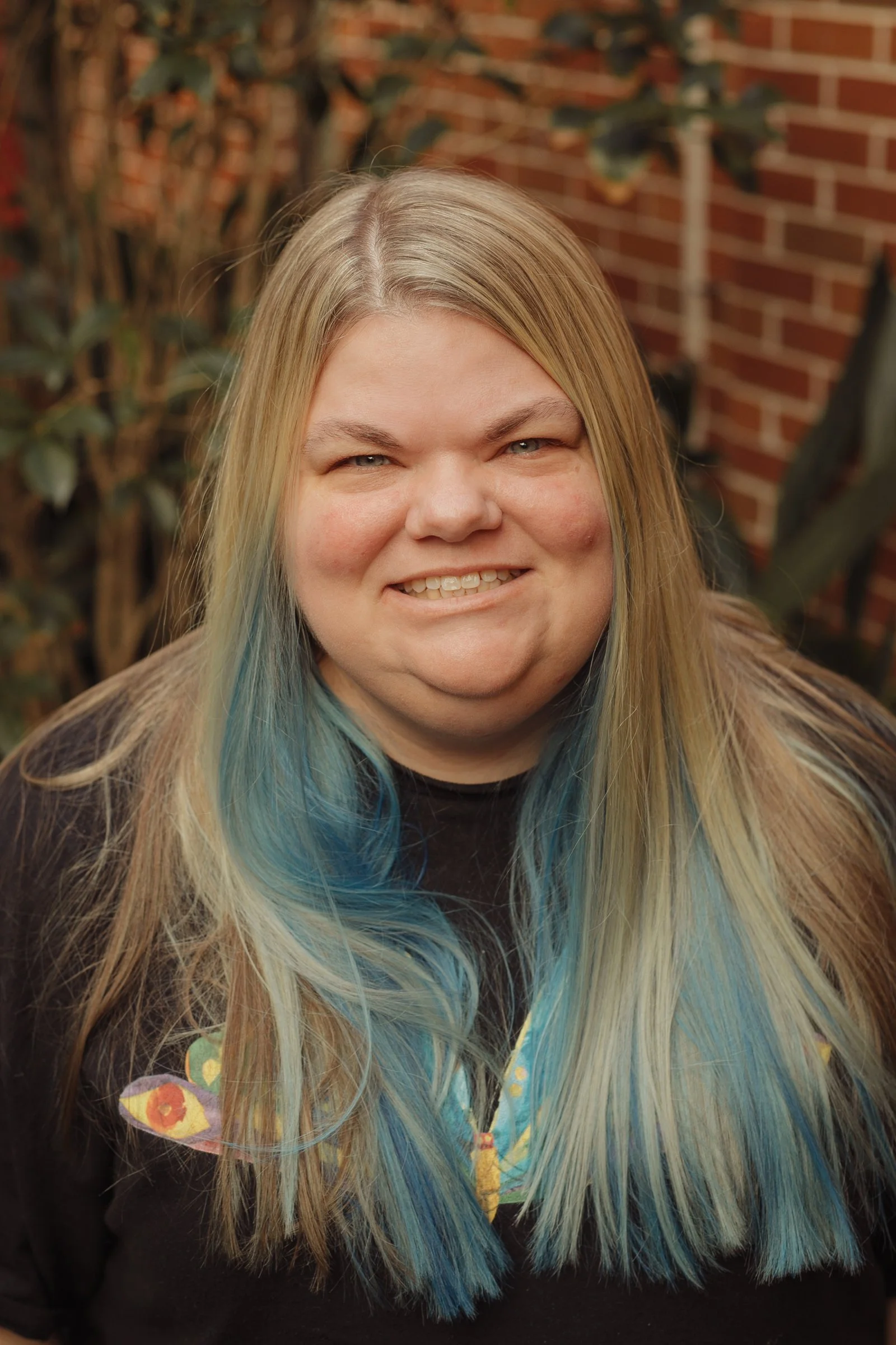 A smiling woman with long blond hair with blue highlights, wearing a black shirt with colorful patches, outdoors near a brick wall and plants.