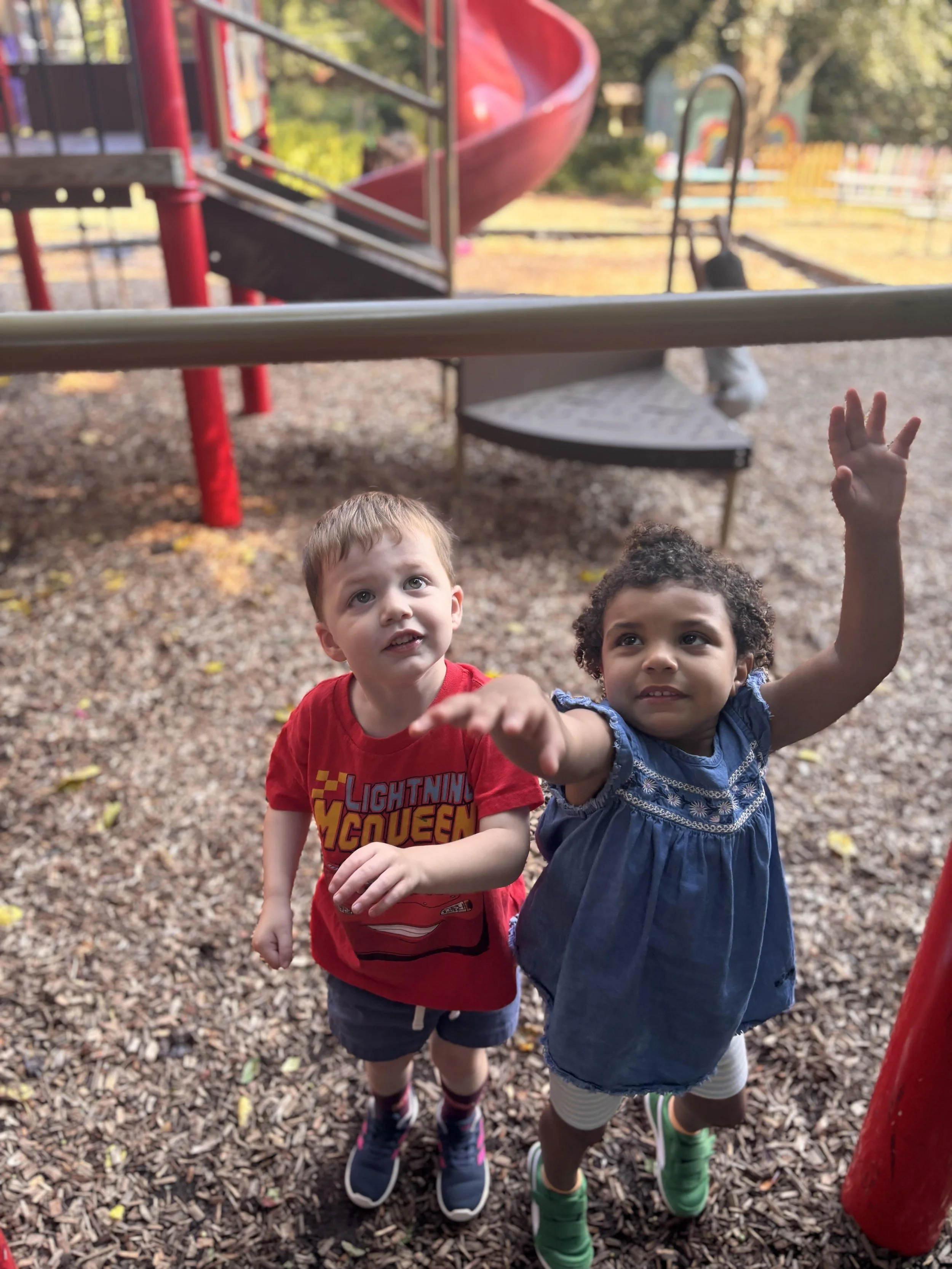 Two young boys standing on a playground filled with wood chips, looking up and pointing, with a red playground slide in the background during daytime.