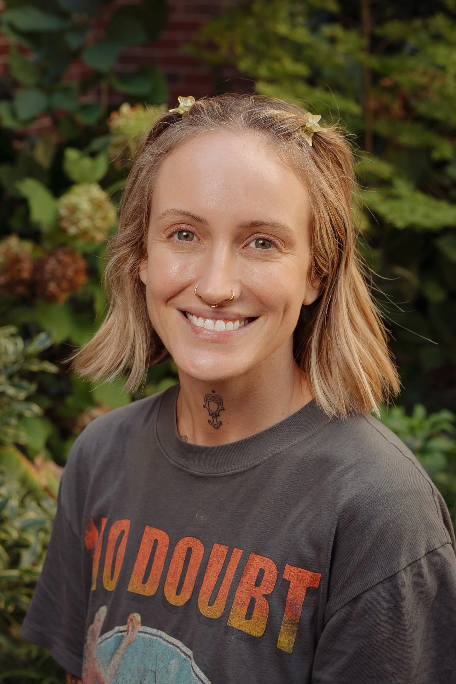 A smiling woman with short blonde hair, nose piercings, tattoos on her neck and chest, wearing a dark T-shirt with the words 'NO DUBT' visible, outdoors with green plants in the background.