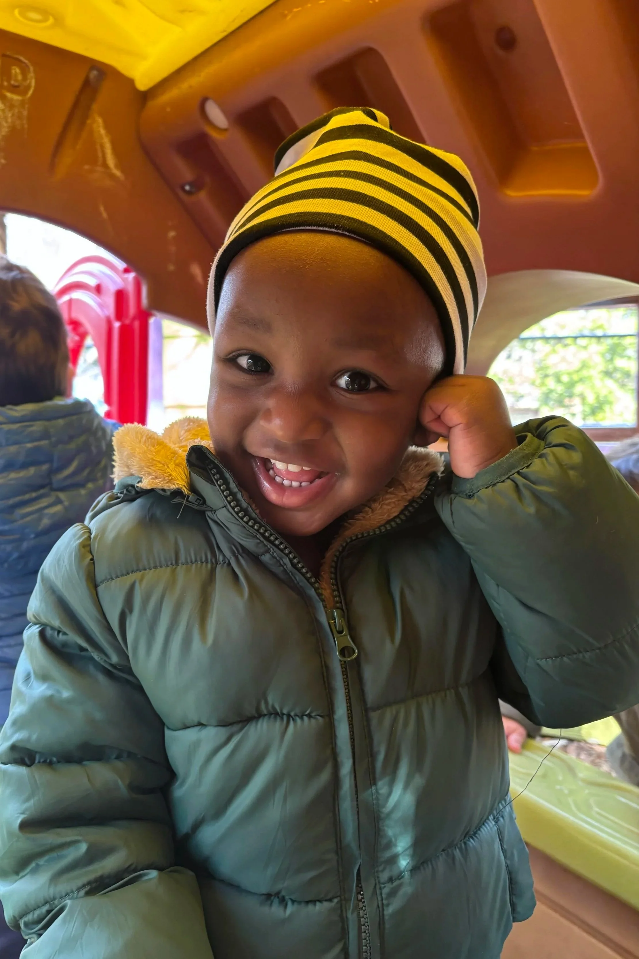 A young boy smiling and wearing a yellow and black striped beanie and a green puffer jacket, inside a playground structure with red, yellow, and green elements.