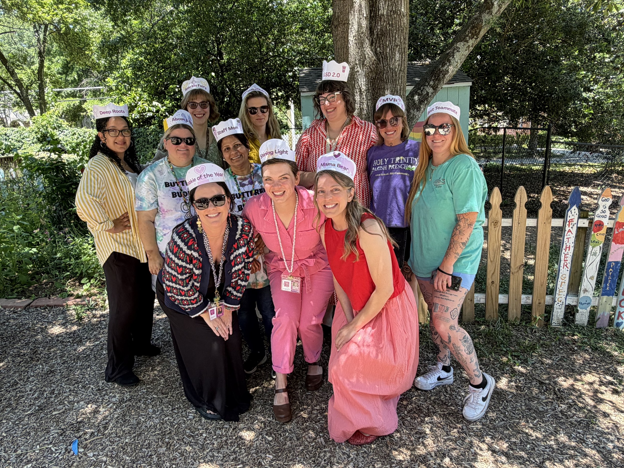 A group of teachers standing outdoors, smiling for a photo in a garden with trees and a small blue shed in the background.