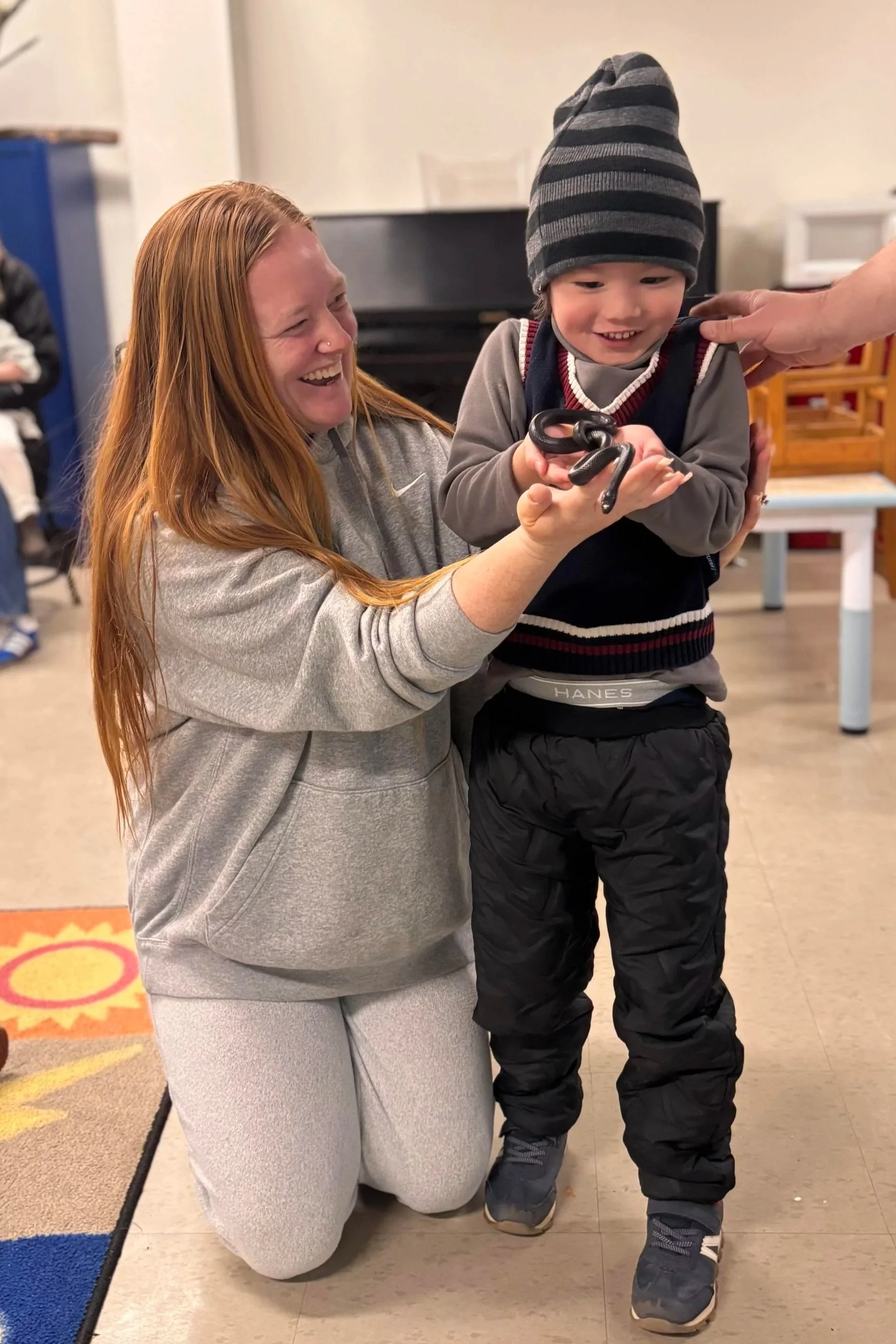 A woman with long red hair, wearing a gray Nike hoodie and light gray sweatpants, kneeling on the floor, smiling at a young boy. The boy, dressed in a striped gray and black beanie, a gray and black jacket, and black snow pants, is holding a black snake in his hand. An adult's hand is gently touching the boy's shoulder. The background shows a room with a piano and some furniture.