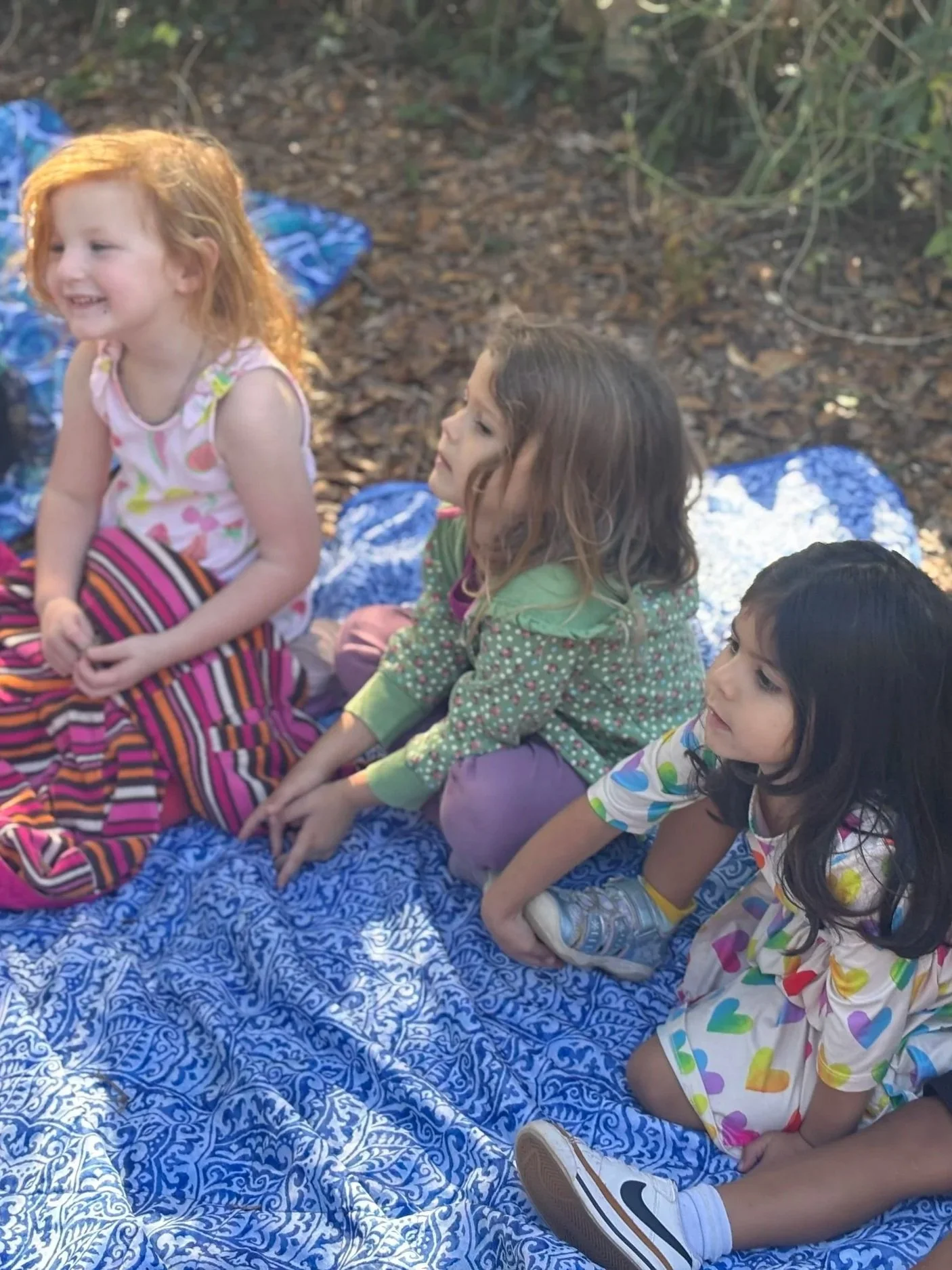 Four young children sitting cross-legged on a blue patterned blanket outdoors among trees, smiling and talking to each other.