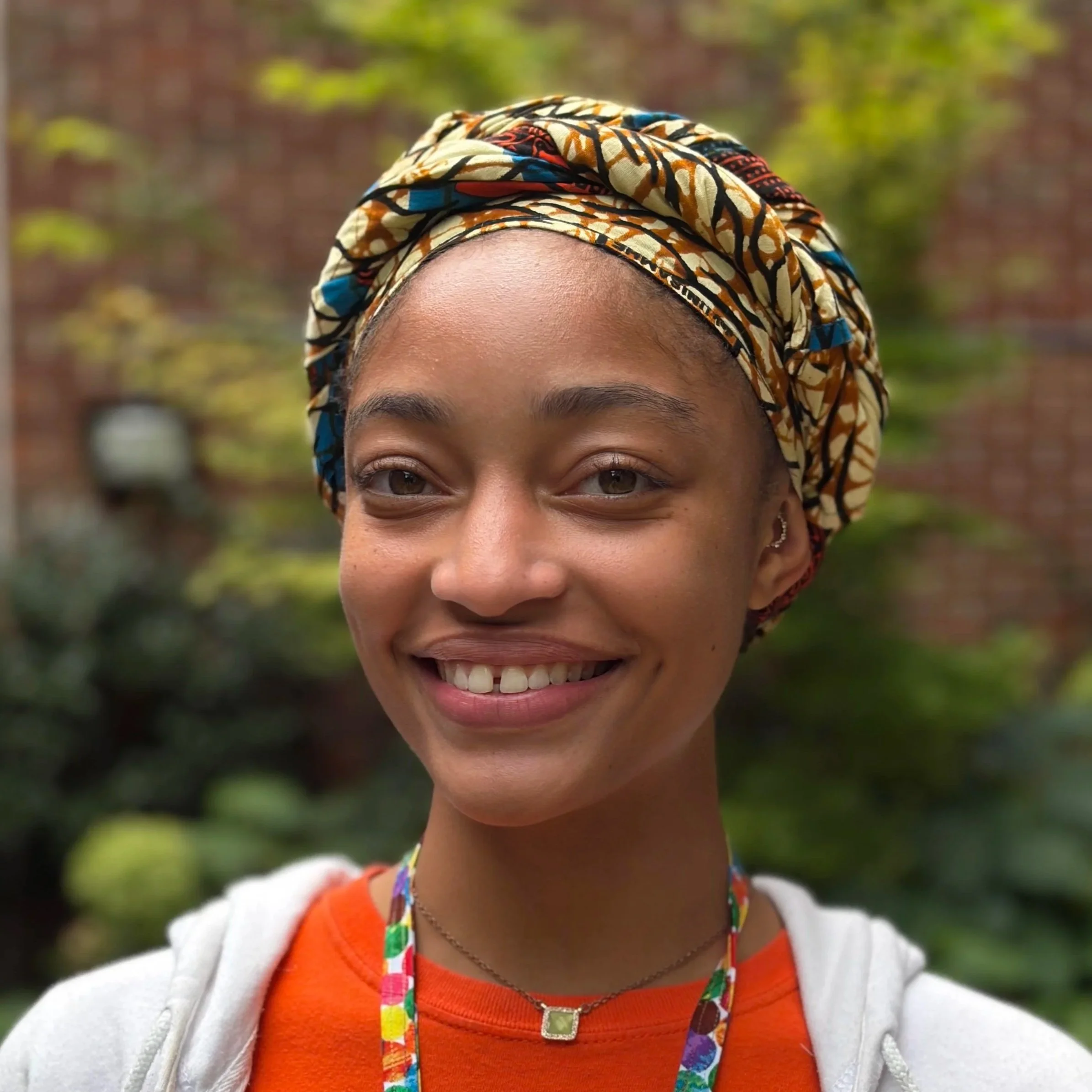 Infinit, a Black woman with dark natural hair, smiles gently in front of a shadowy outdoor setting. She wears a yellow turtleneck and their head is tilted to one side. 