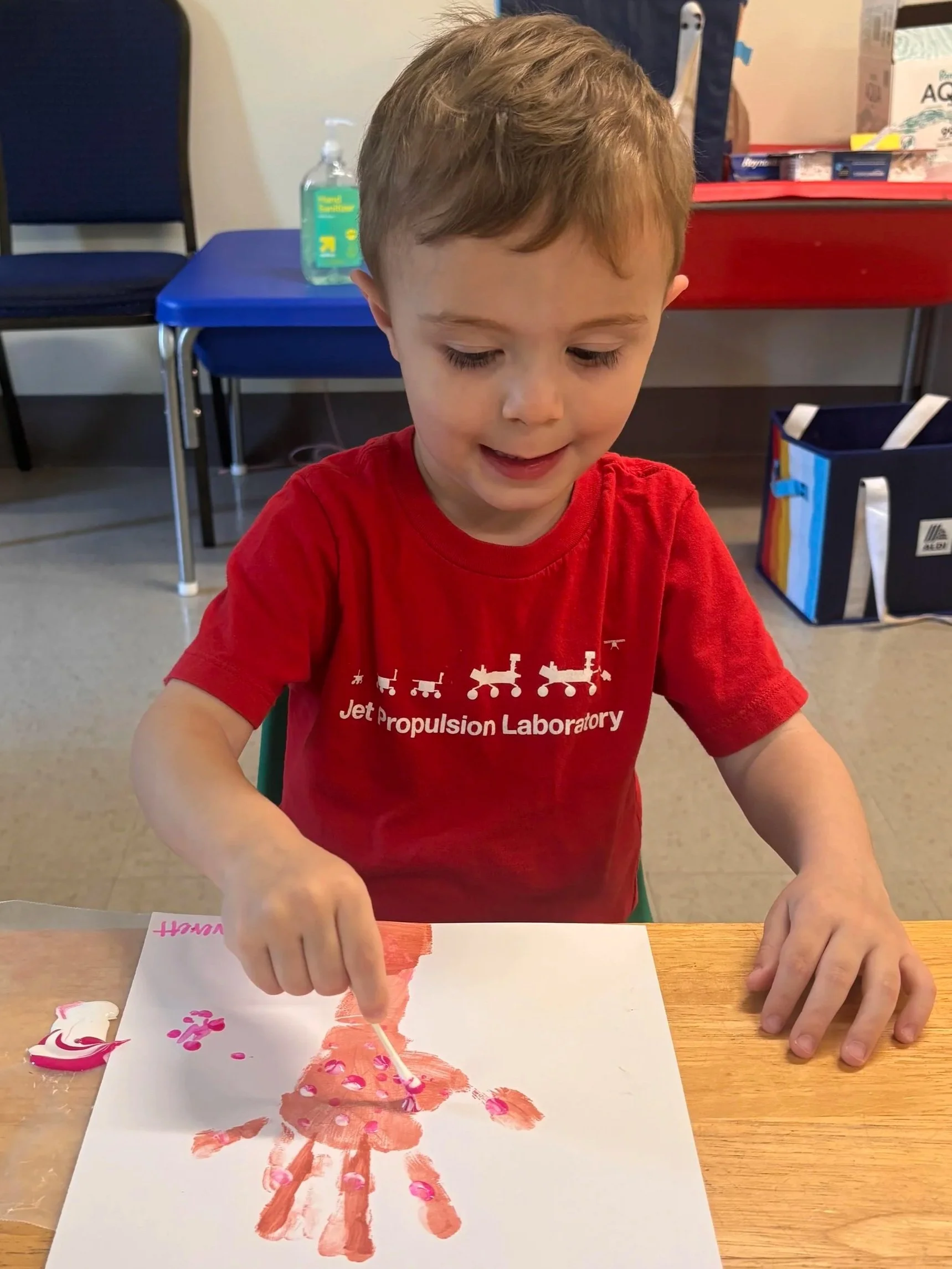 A young boy in a red T-shirt is painting a picture on a piece of paper at a table. He is using a paintbrush to apply pink paint on the paper, creating a floral or abstract design.