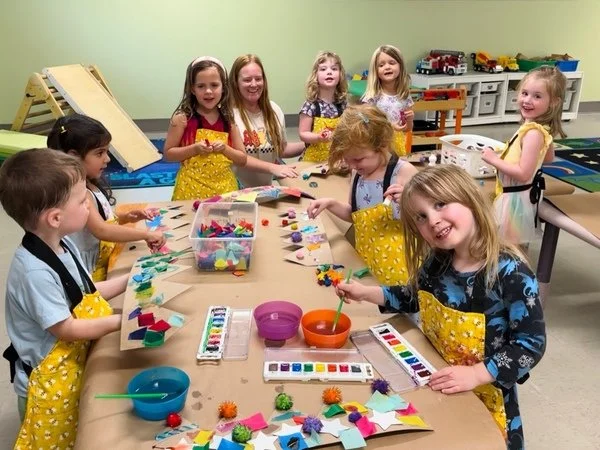 Children in a craft classroom working on colorful felt projects, wearing aprons, with craft supplies on the table.