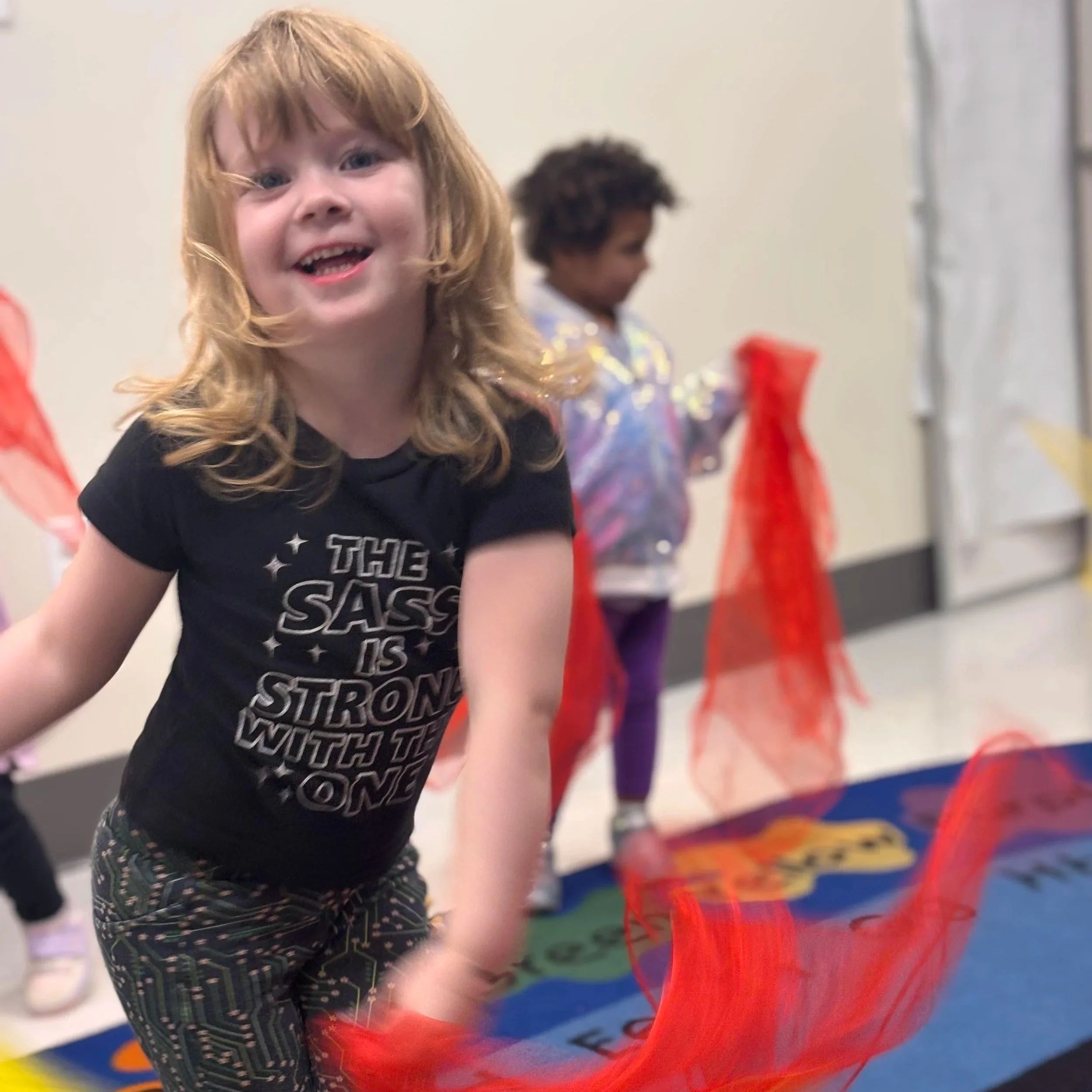 Young girl smiling and playing with orange fabric in a classroom. Other children are in the background.