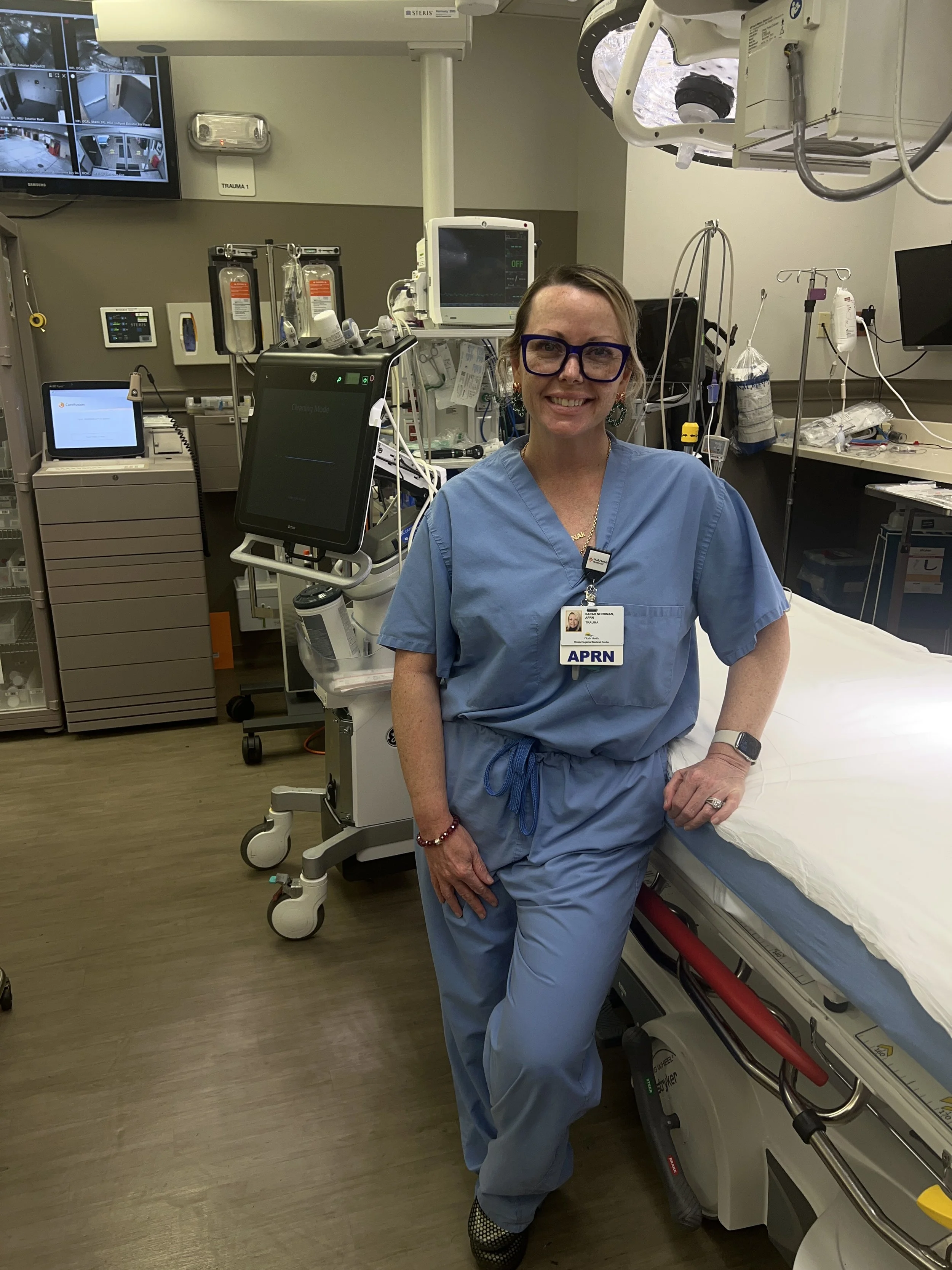 A smiling female healthcare worker in blue scrubs standing next to a hospital bed in a hospital room filled with medical equipment and monitors.