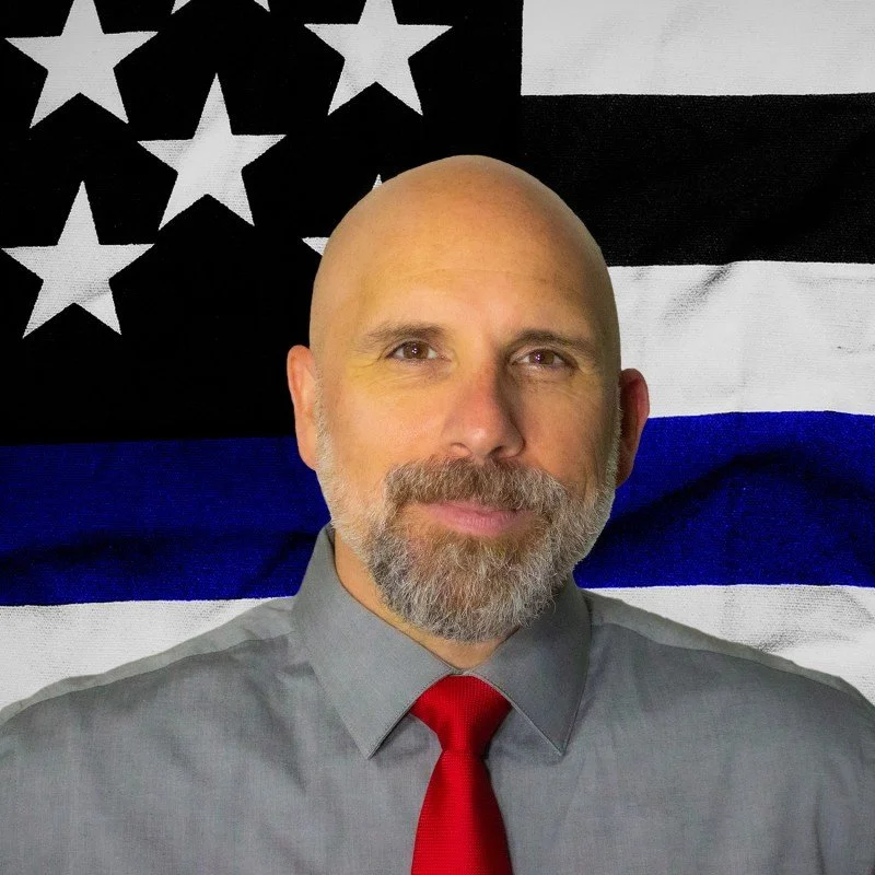 A man with a shaved head and beard wearing a gray shirt and red tie standing in front of a black, white, and blue American flag with a thin blue line.