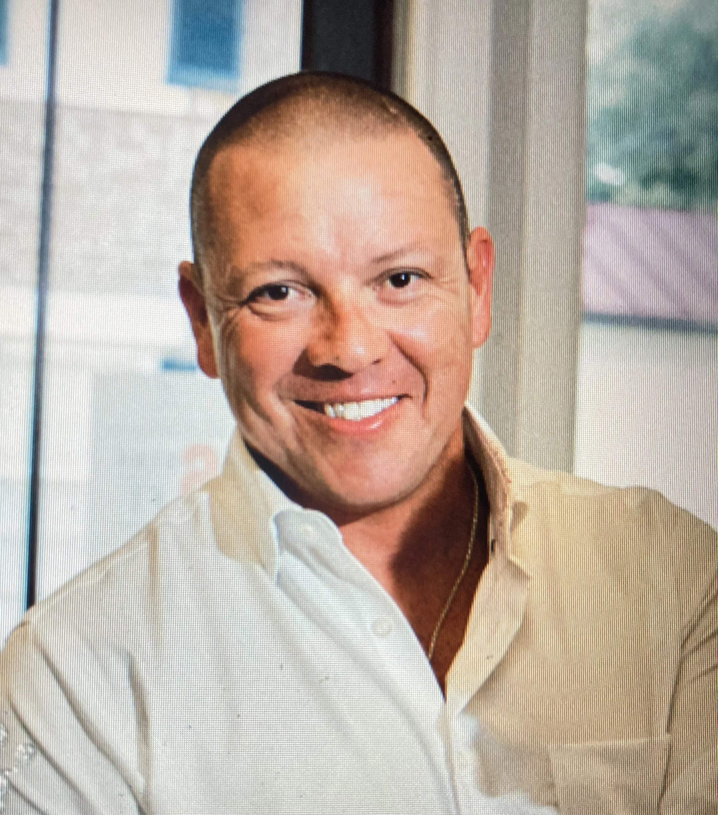 A smiling man with a shaved head, wearing a white collared shirt, standing indoors near a window.