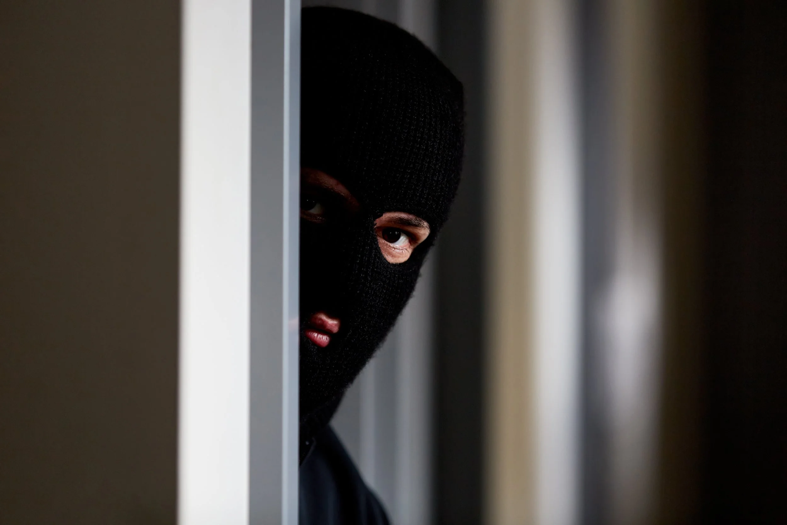 Person wearing a black ski mask peering out from behind a gray vertical surface.