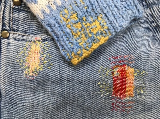 Veste en denim avec deux broderies de bougies allumées, une en haut à gauche et une en bas à droite, accompagnées de rayons de lumière.