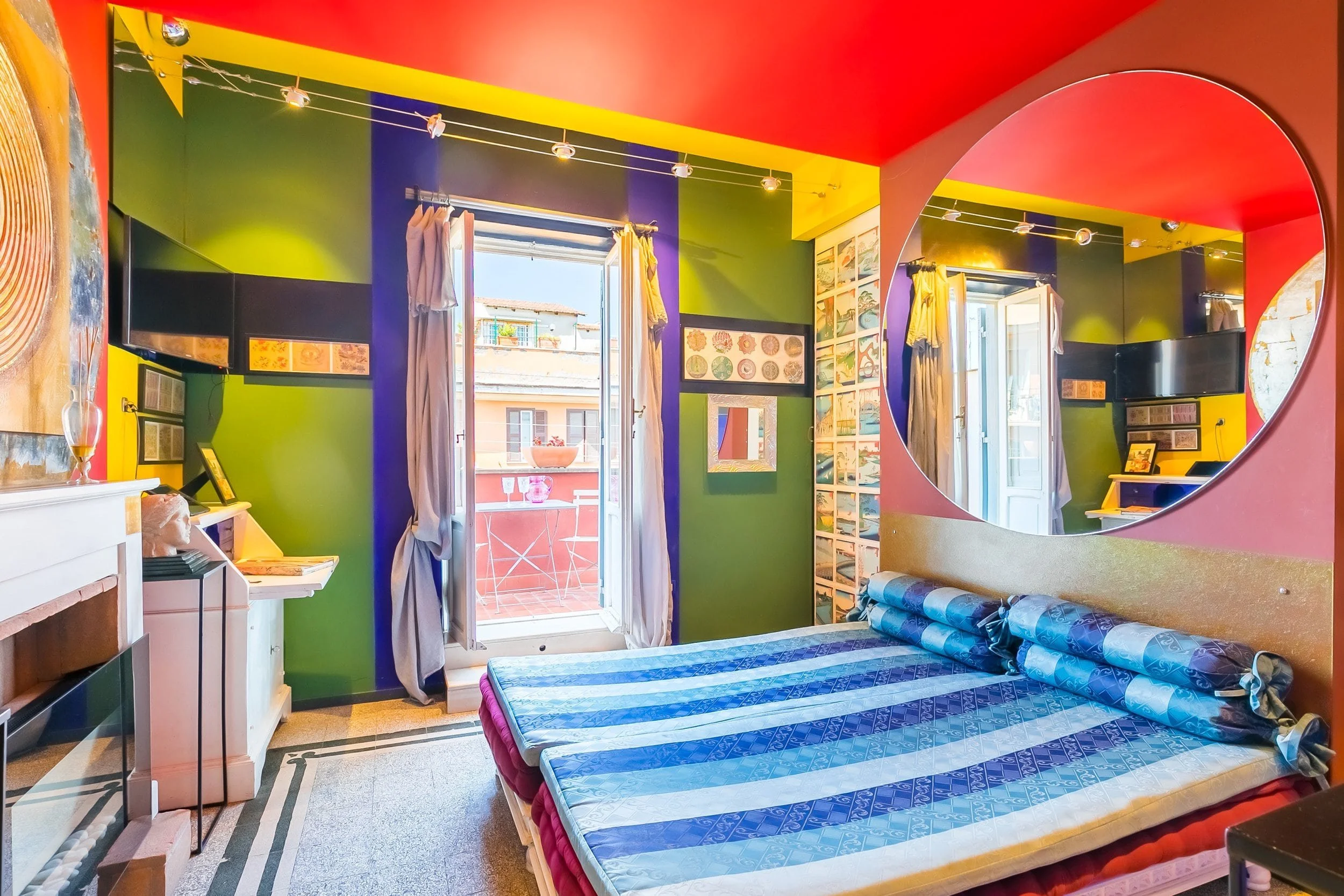 Colorful bedroom with green, purple, and red walls, a double bed with blue patterned bedding, a large round mirror on the wall, an open balcony door with a small table and two chairs outside, and various wall decorations.