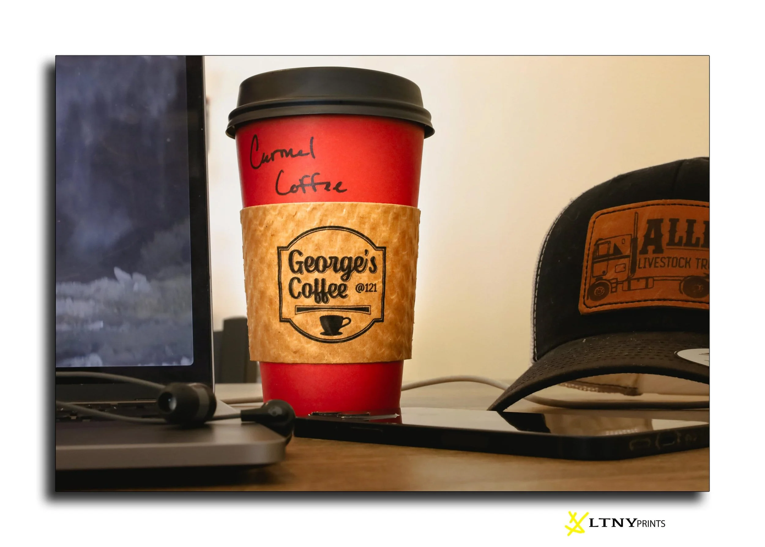 A red to-go coffee cup with a black lid, labeled 'Carmel Coffee' handwritten on it, and a brown sleeve that reads 'George's Coffee' with a small coffee cup icon. The cup is on a desk next to a laptop, a cap with an embroidered palm tree and sunset, and a pair of black earbuds connected to the laptop.