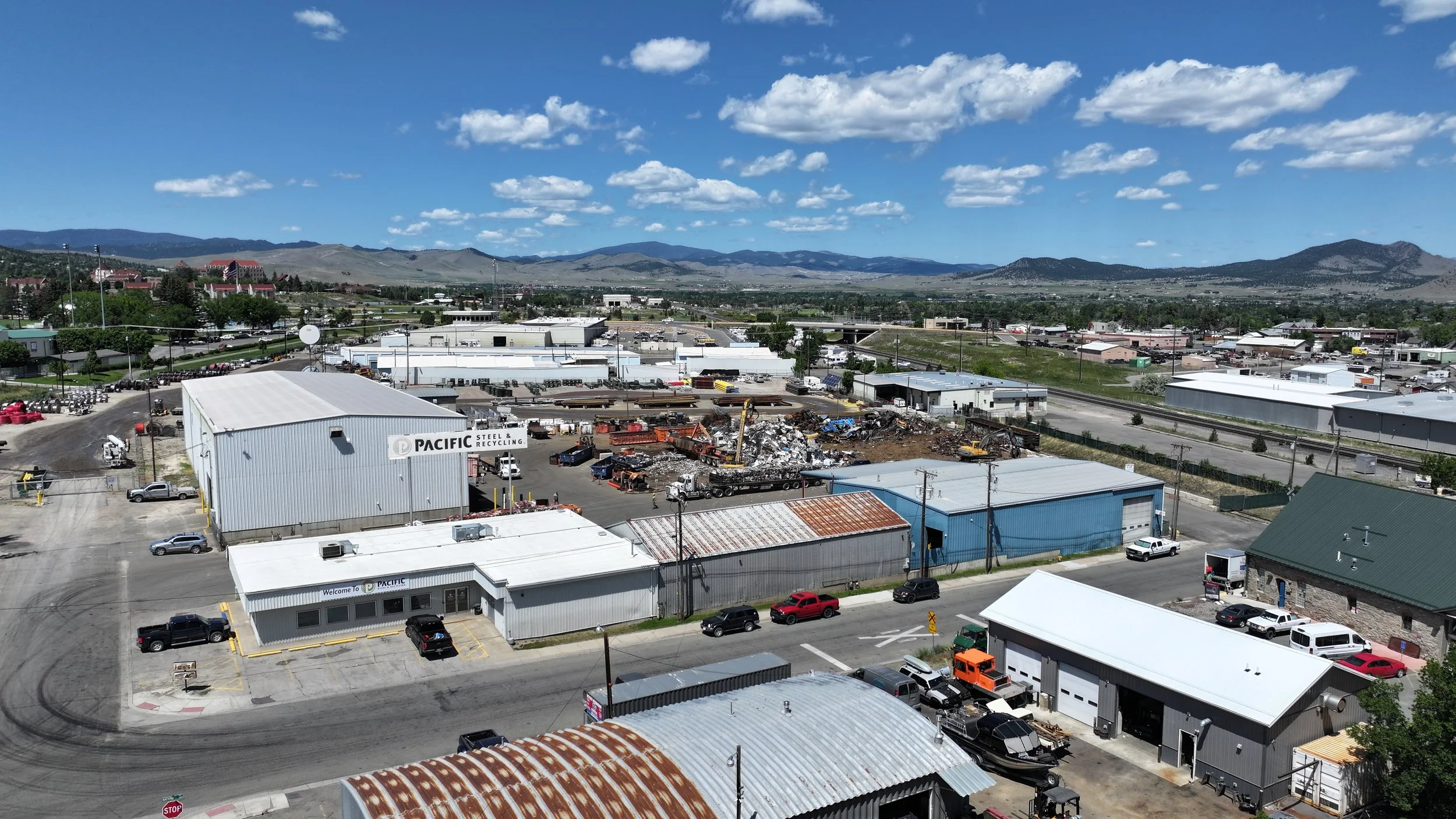 Aerial view of an industrial area with warehouses, parking lots, and a scrap yard with debris and machinery, with mountains in the background under a partly cloudy sky.
