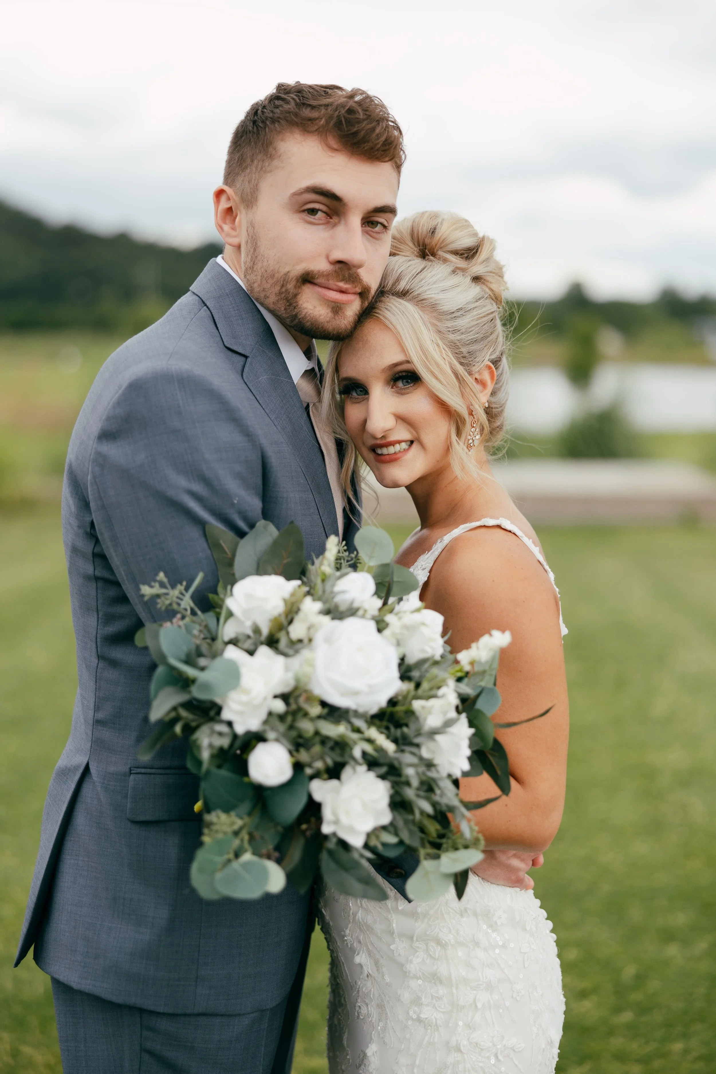Bride and groom embracing outdoors, groom in gray suit, holding bouquet with white flowers and greenery.