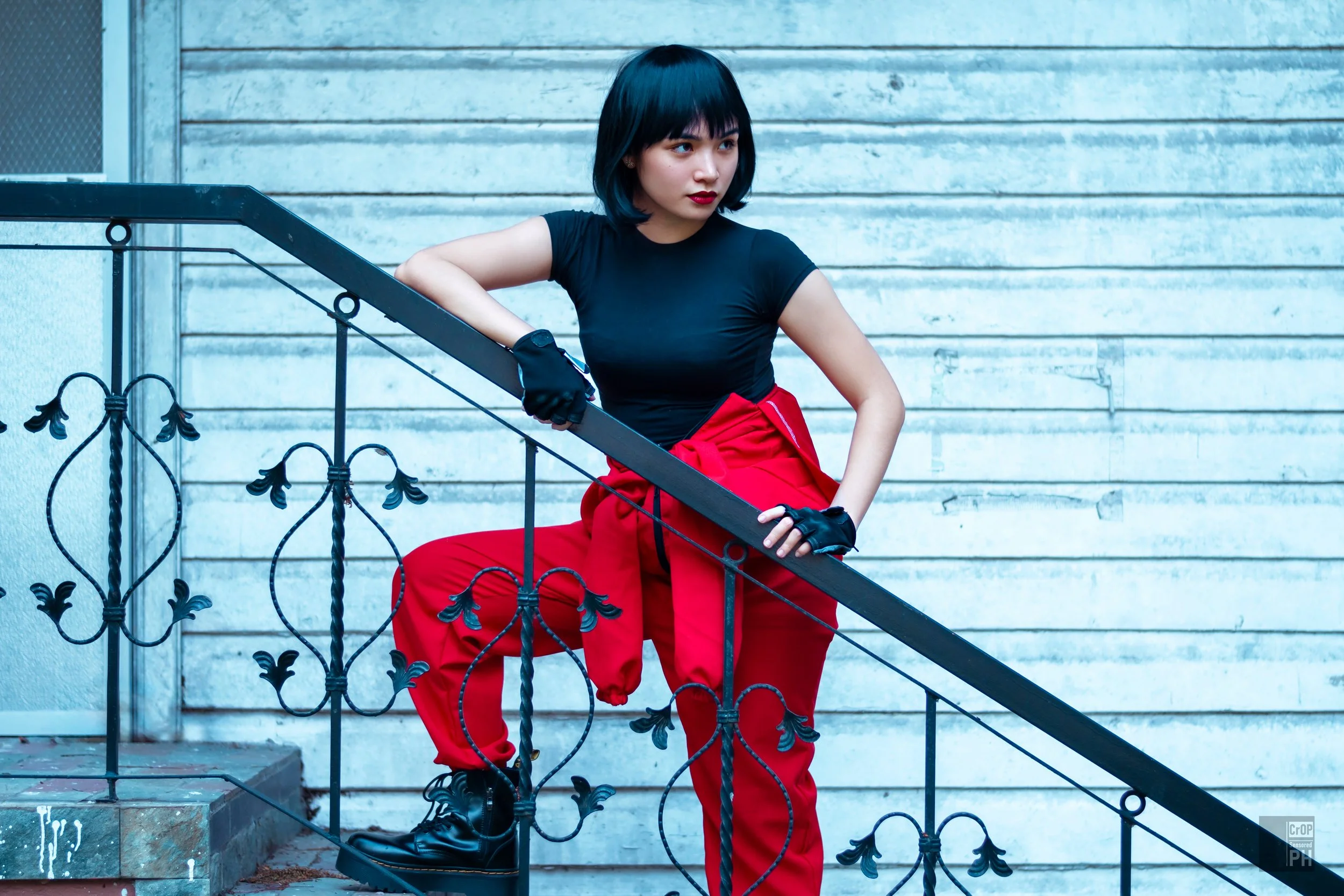 Young woman with black hair, red lipstick, wearing a black t-shirt, red pants, and gloves, sitting on a decorative black iron staircase outside against a concrete wall.
