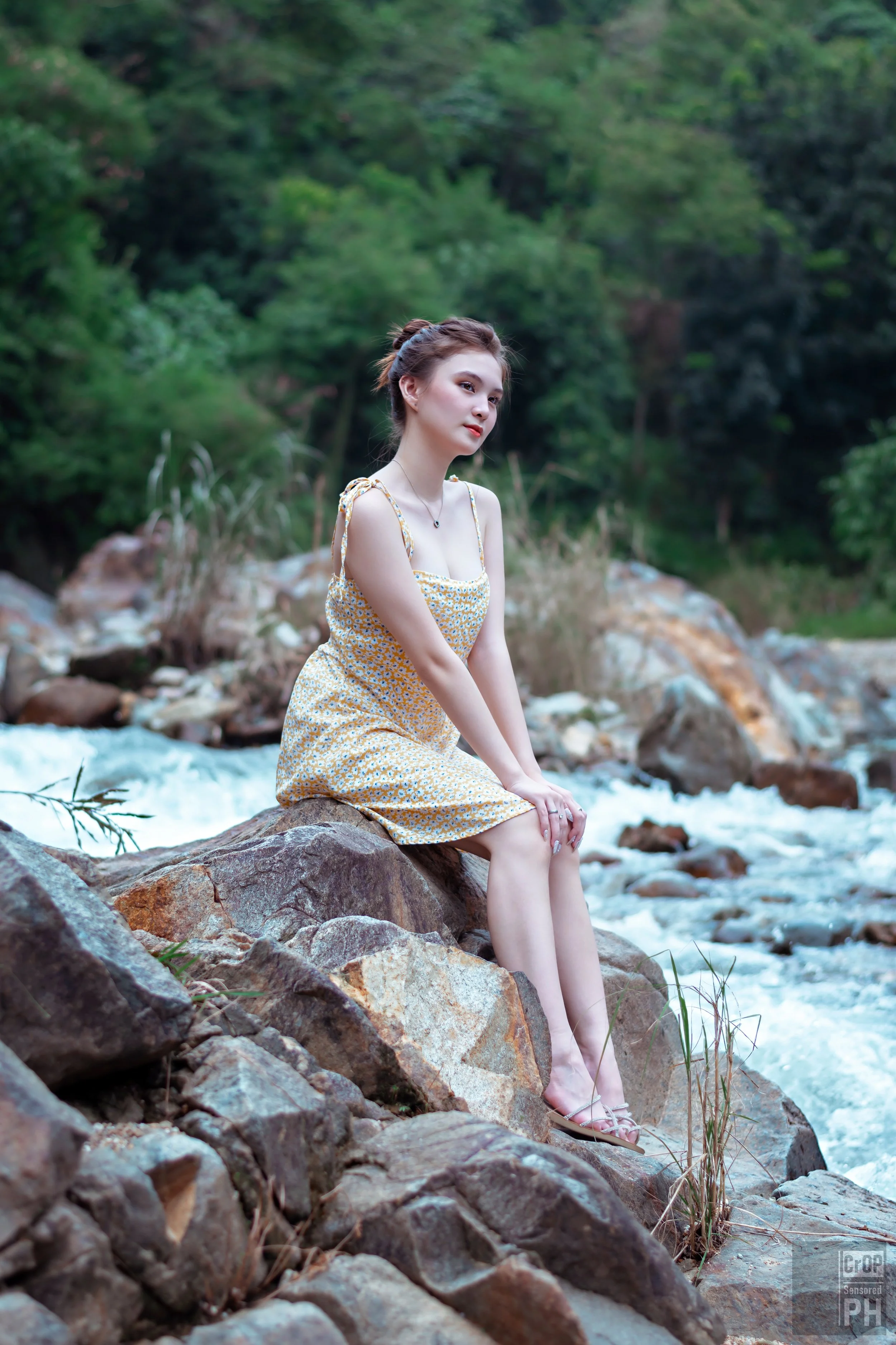 A young woman in a yellow floral dress sits on a rock by a flowing stream, surrounded by lush green trees in a wooded area.