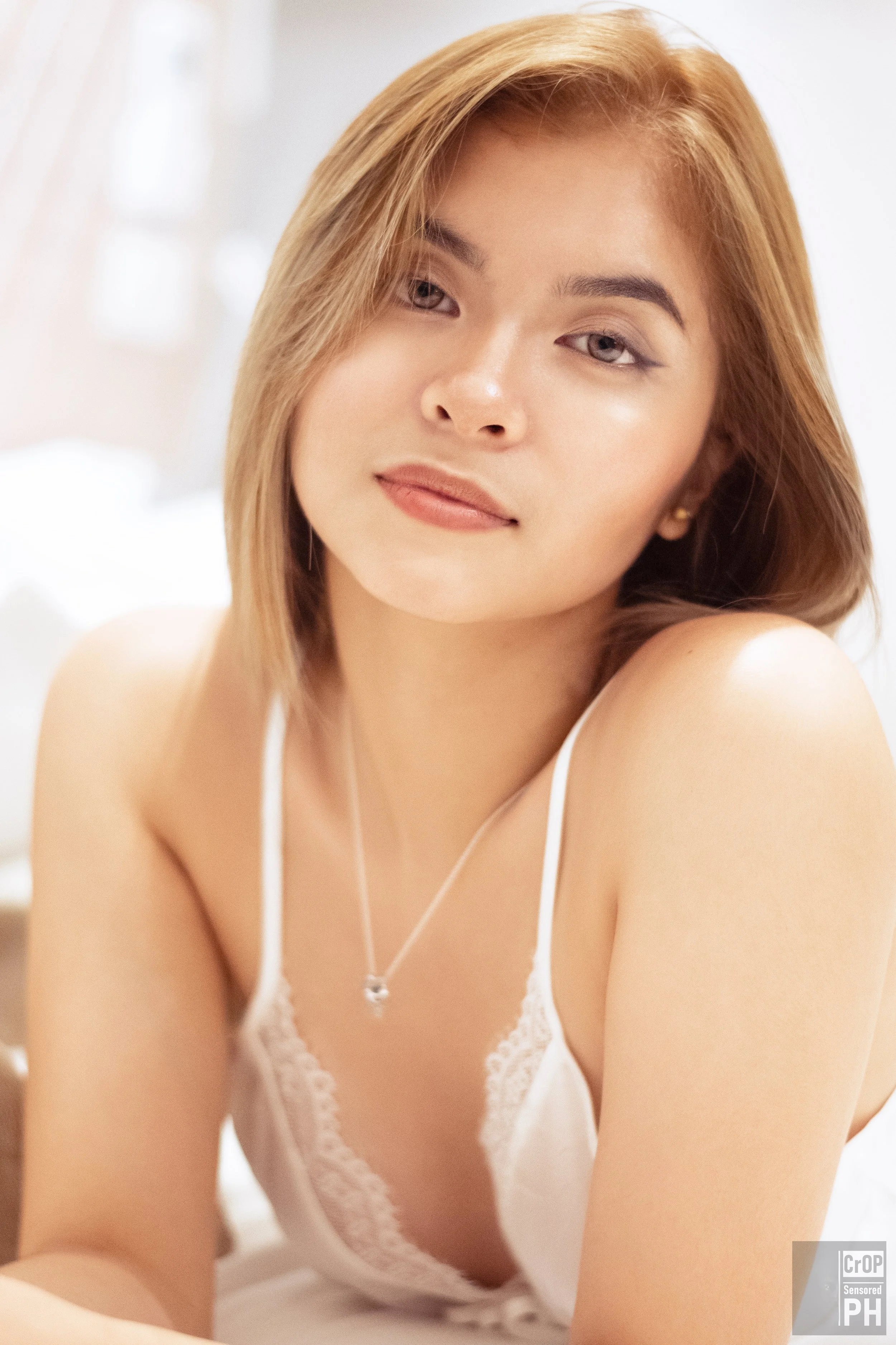 A young woman with blonde hair and light blue eyes, wearing a white lace camisole, sitting on her knees with a soft smile in a bright, softly blurred background.