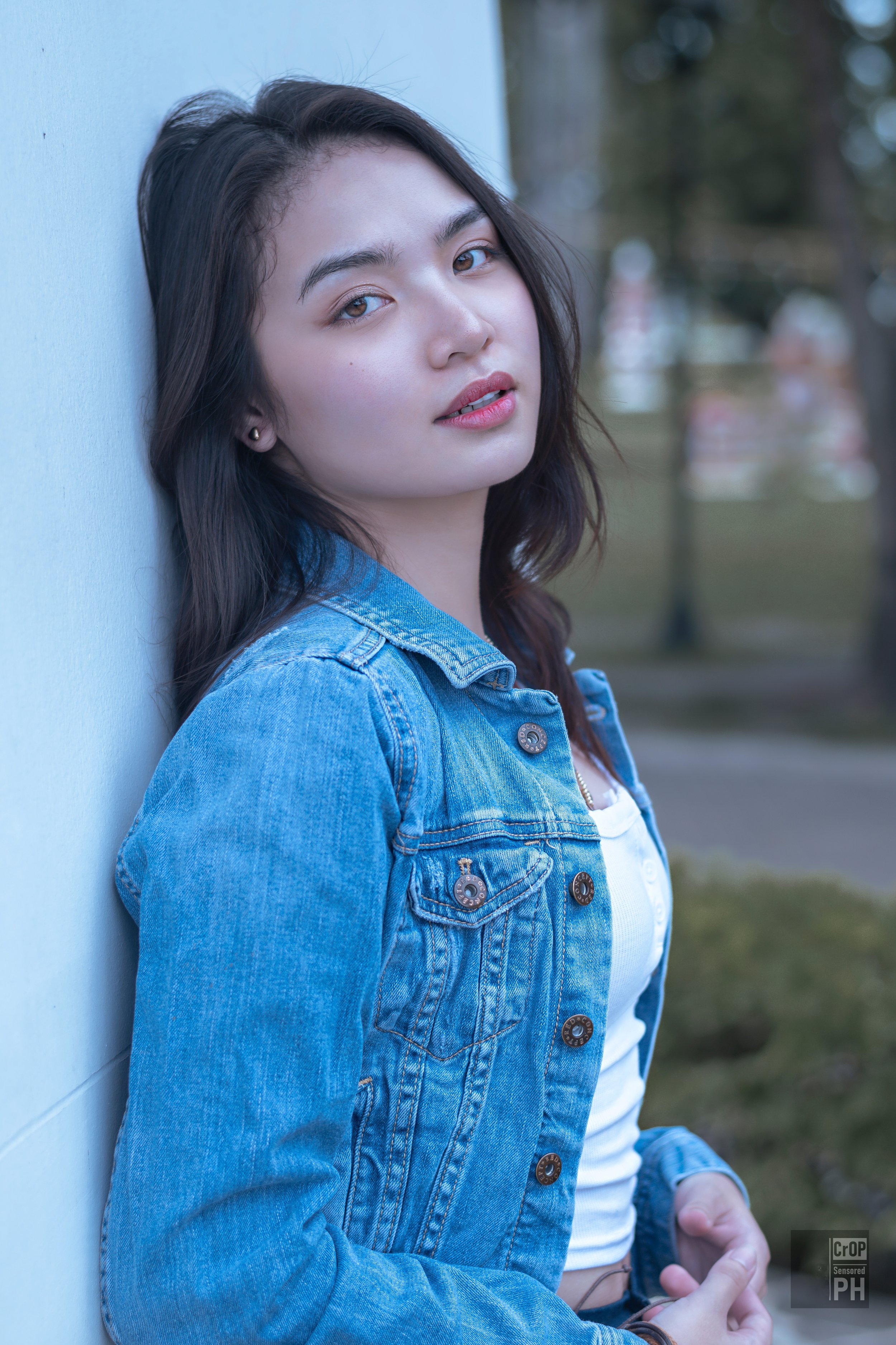 A young woman with dark hair and light skin, wearing a denim jacket and a white top, leaning against a blue wall outdoors. She has light makeup and is looking at the camera with a slight smile.