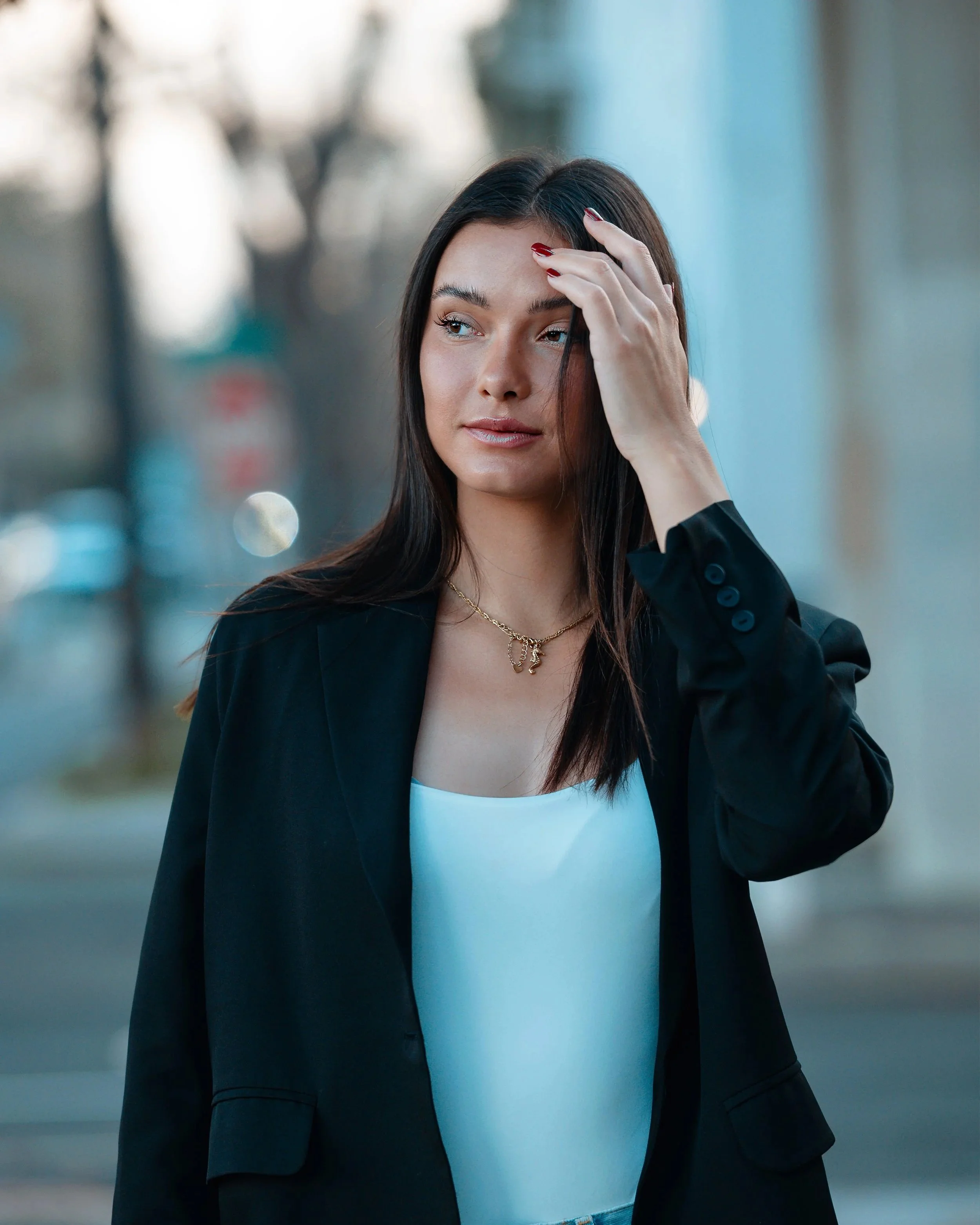 A woman with long dark hair, wearing a black blazer, white top, gold necklace, and red nail polish, standing outdoors, touching her forehead and looking to the side.