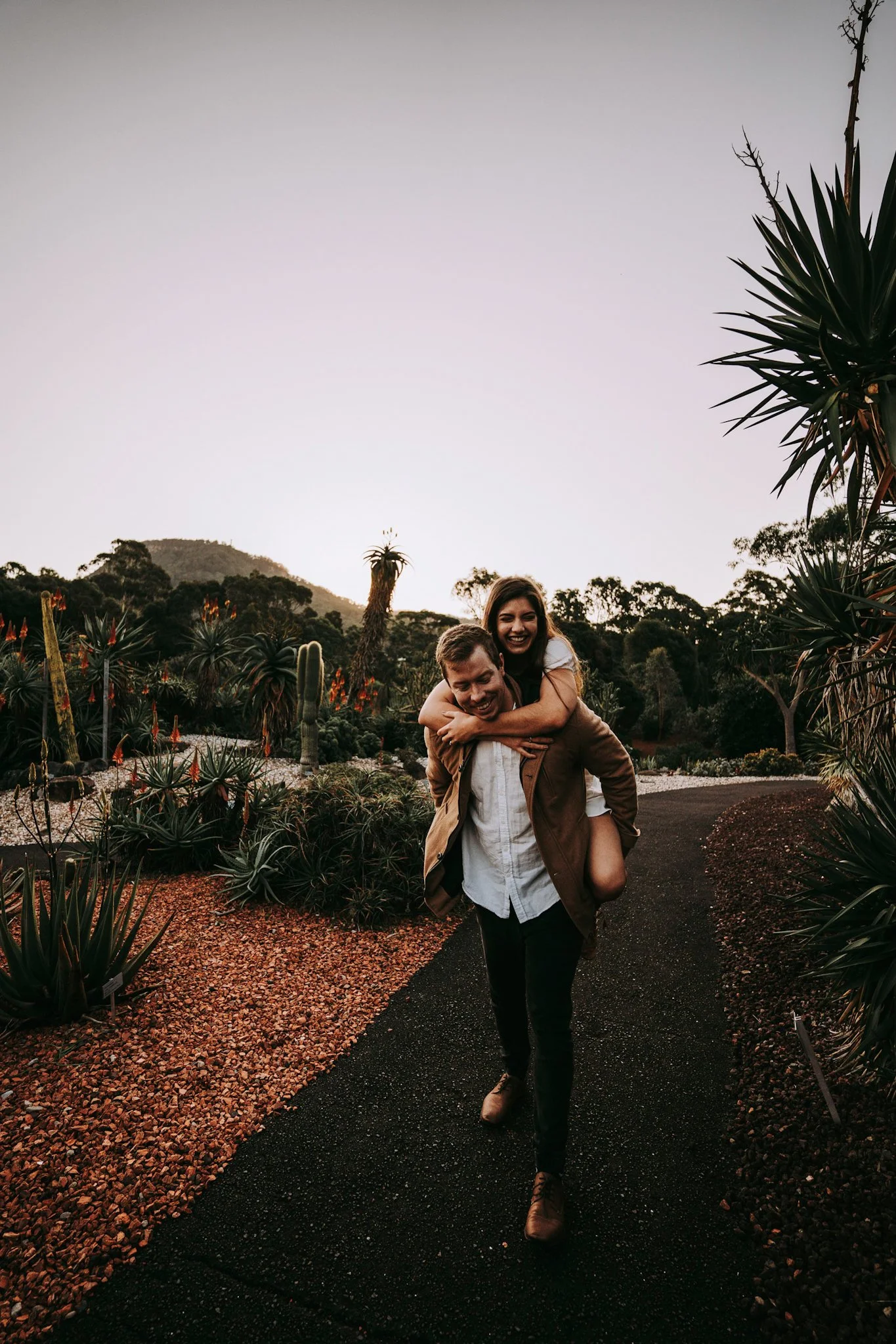 A happy couple, man carrying woman on his back, walking on a garden path at sunset with verdant plants and mountains in the background.