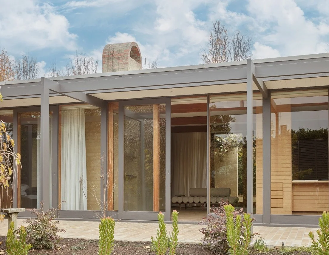 Modern house with large glass windows and a patio, surrounded by a garden with plants and shrubs.