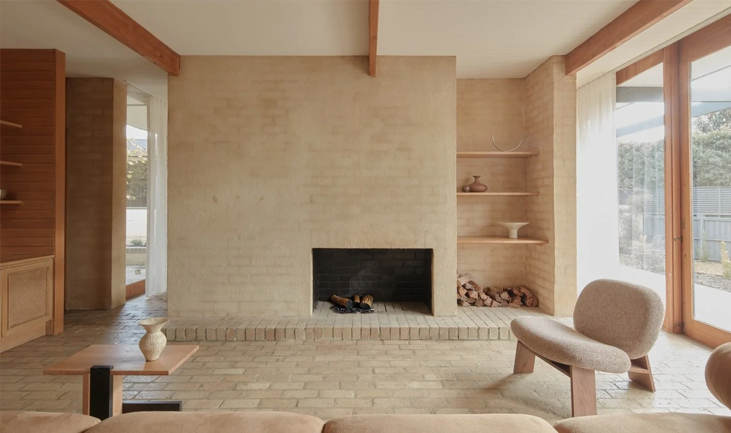 Living room with a brick fireplace, beige brick walls, large windows with curtains, and wooden beams on the ceiling. There is a small side table with a decorative object, a modern chair, and built-in shelves with decorative items and firewood.
