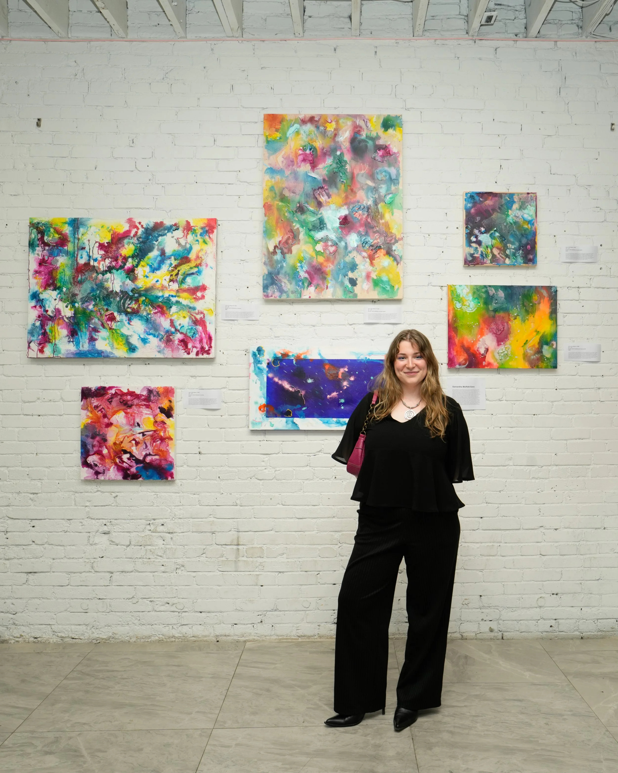 The artist, with her hair down, wearing all black, witchy boots & a vintage pink purse, standing in front of her Ink to the Abyss series. The colourful, textured abstract ink works are displayed on a white painted brick wall