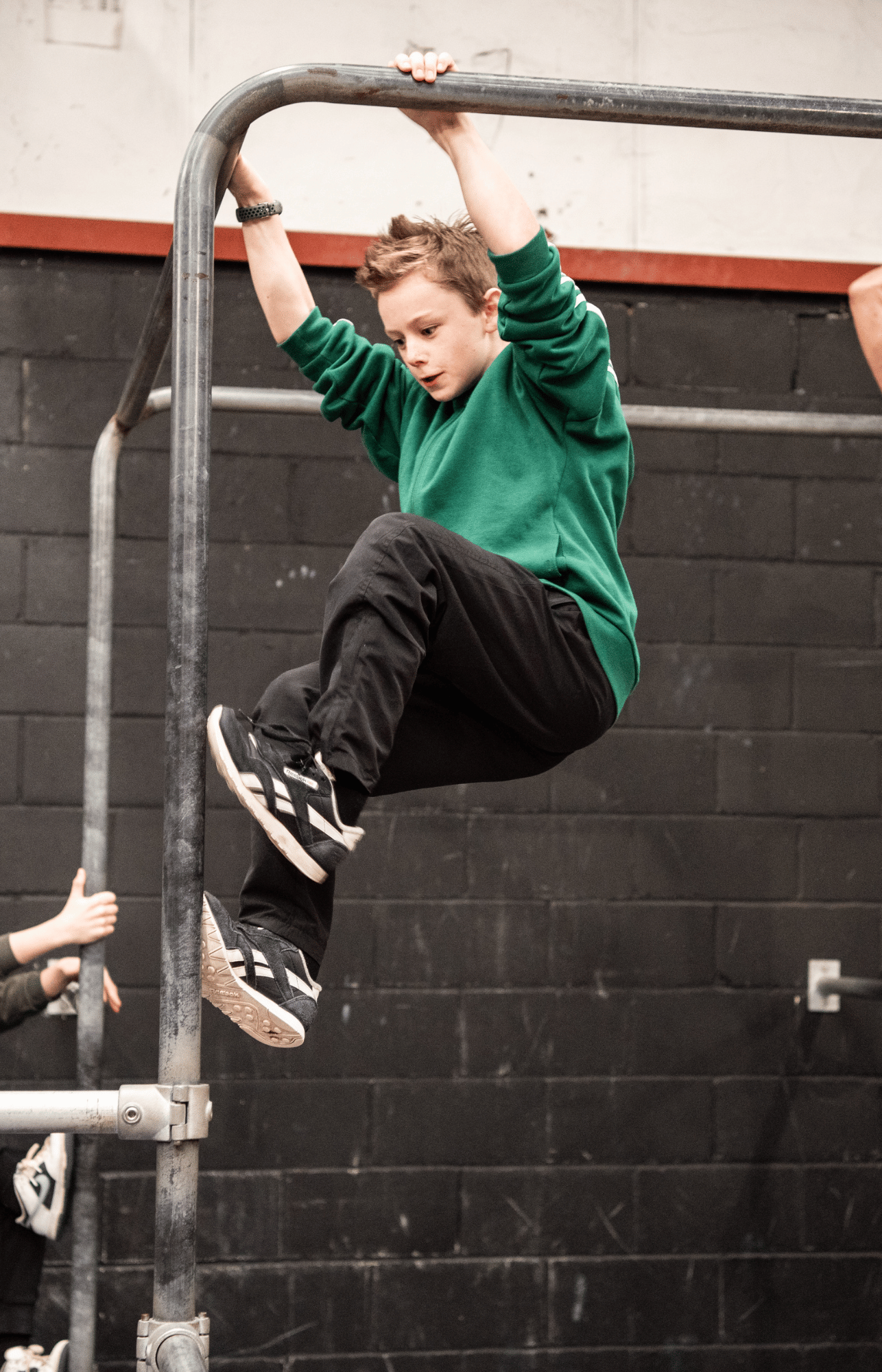 Parkour and Free Running student climbing scaffolding bar in green top and reebok classics at a parkour gym in Crawley West Sussex in an article about managing fear and resilience.  Child is in cat hang position ready to attempt pole climb up