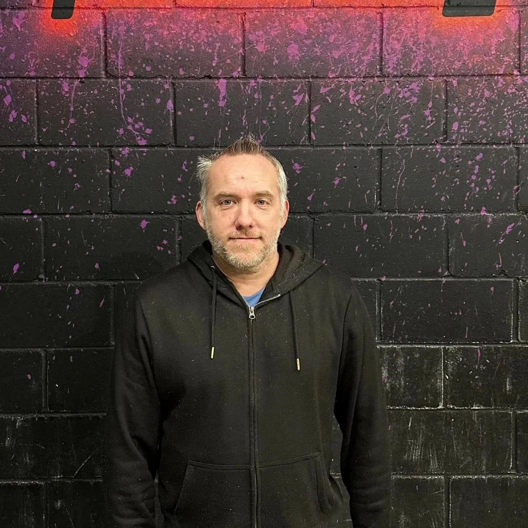 A man with light hair and a beard standing in front of a black and purple splatter painted wall, wearing a black hoodie.